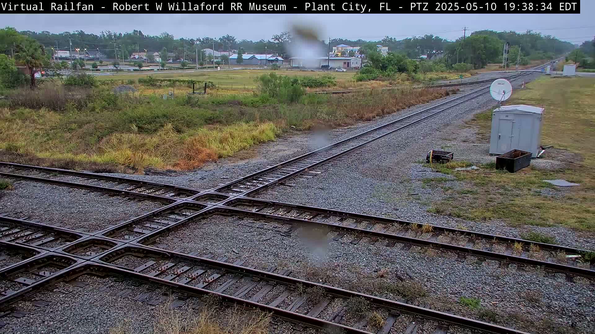 A railroad crossing with multiple tracks is shown in a lightly raining outdoor setting, near some buildings and sparse vegetation.