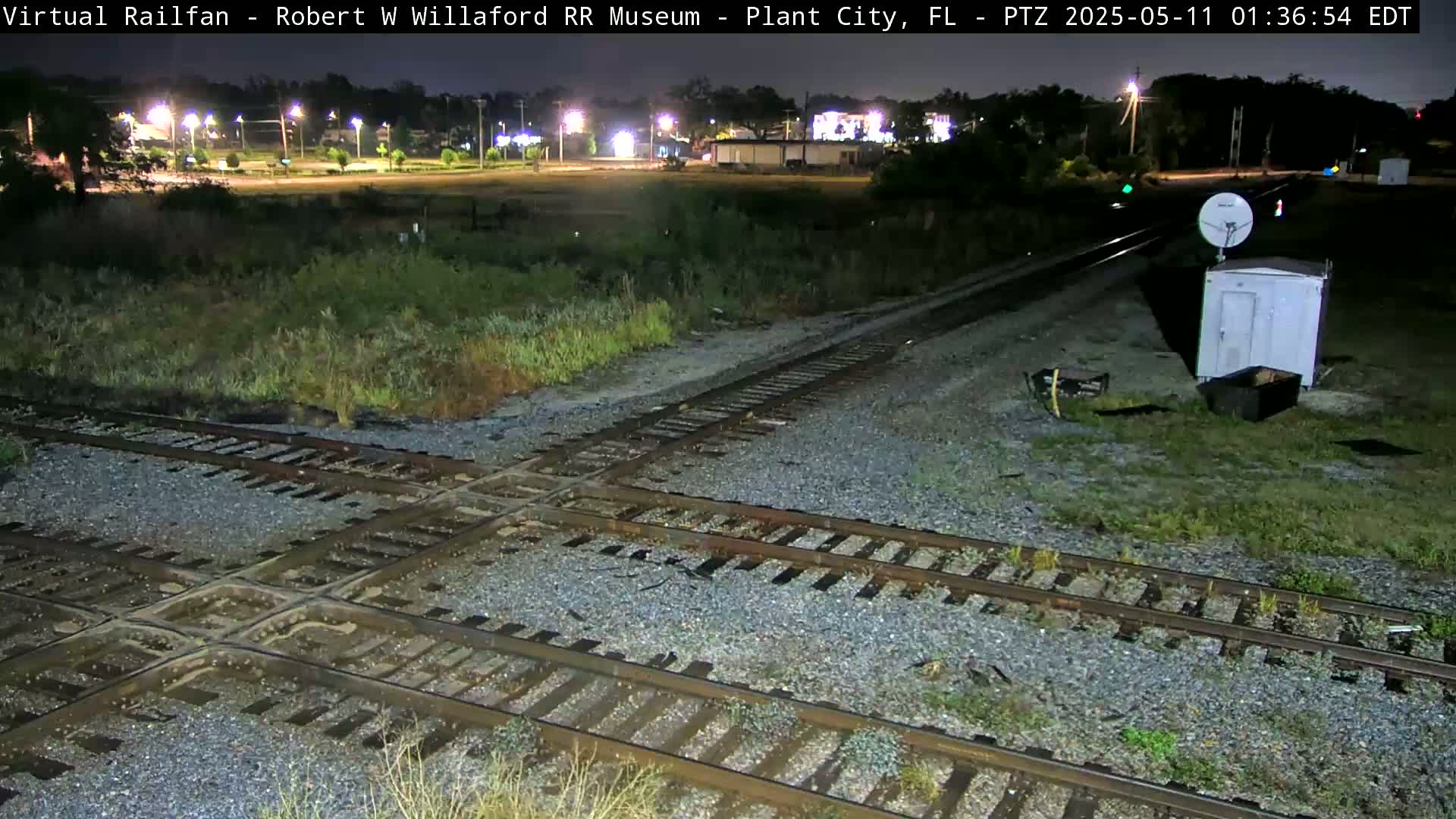 A nighttime view of a railroad crossing with a small building and satellite dish, under dark skies.