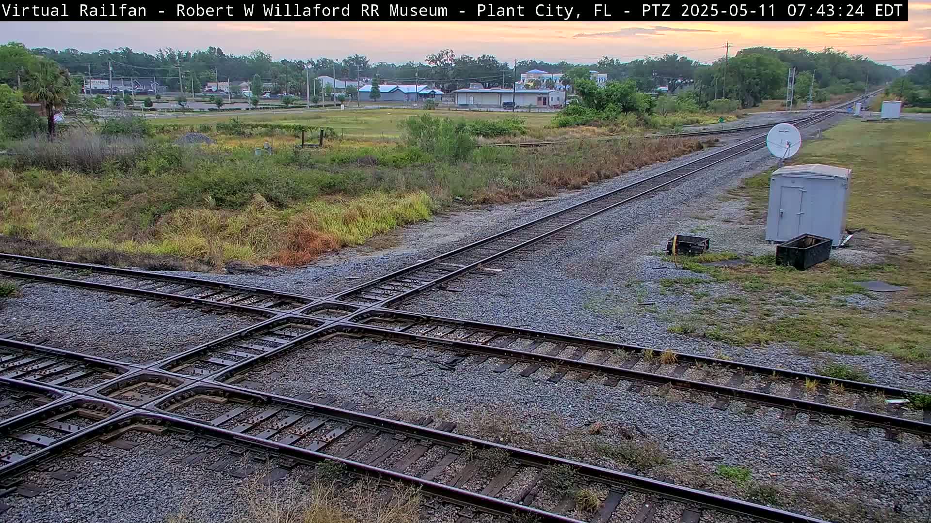 A railroad crossing is shown under a partly cloudy sky at dawn, with a small utility shed and satellite dish nearby.