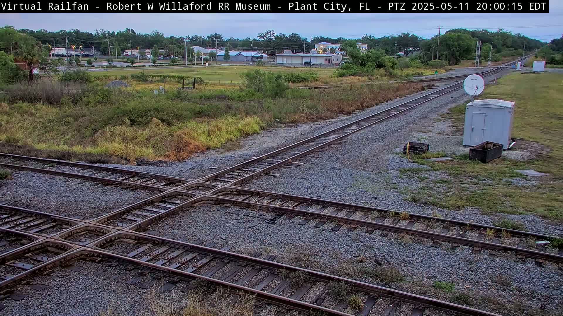 A railroad crossing with intersecting tracks is shown under a twilight sky, next to a small building and satellite dish.