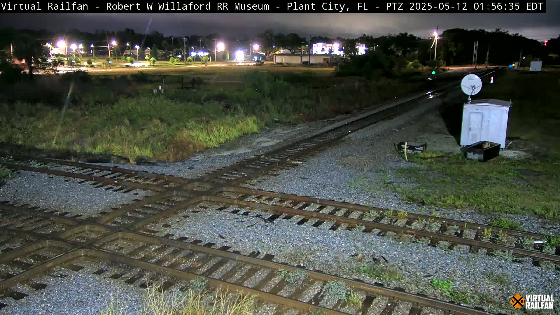 A nighttime view of a railroad crossing with wet tracks and a small building nearby under overcast skies.