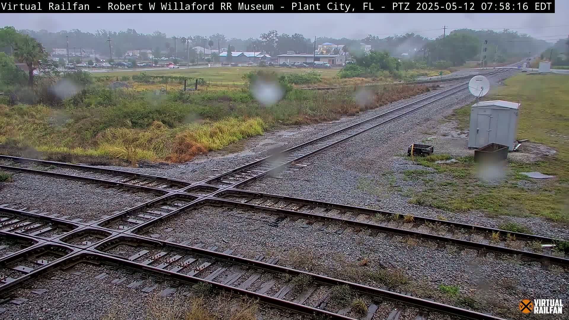 A rainy view of a railroad crossing with a small building and satellite dish nearby, showing a section of track in the foreground and a distant view of buildings and sparse vegetation.