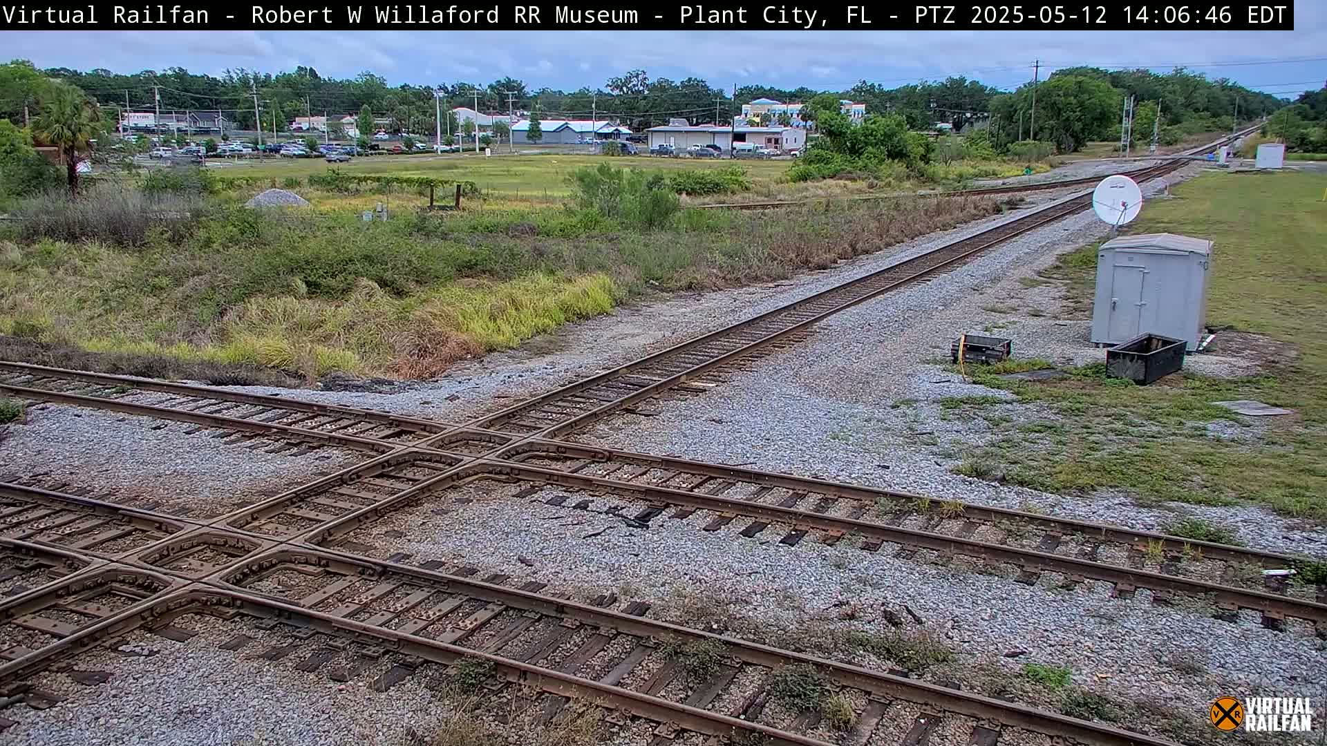 A railroad crossing with several tracks intersects amidst a grassy area, under a partly cloudy sky, with buildings visible in the distance.