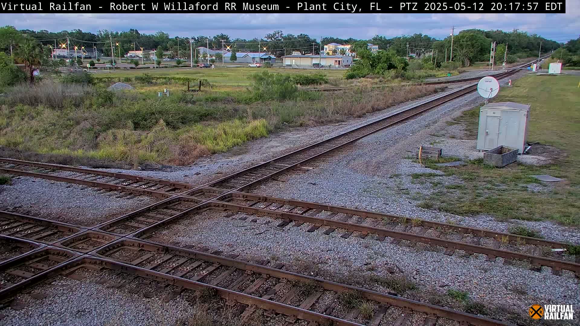 A railroad crossing with two sets of tracks intersects under an overcast sky, with low vegetation and a small building visible in the background.