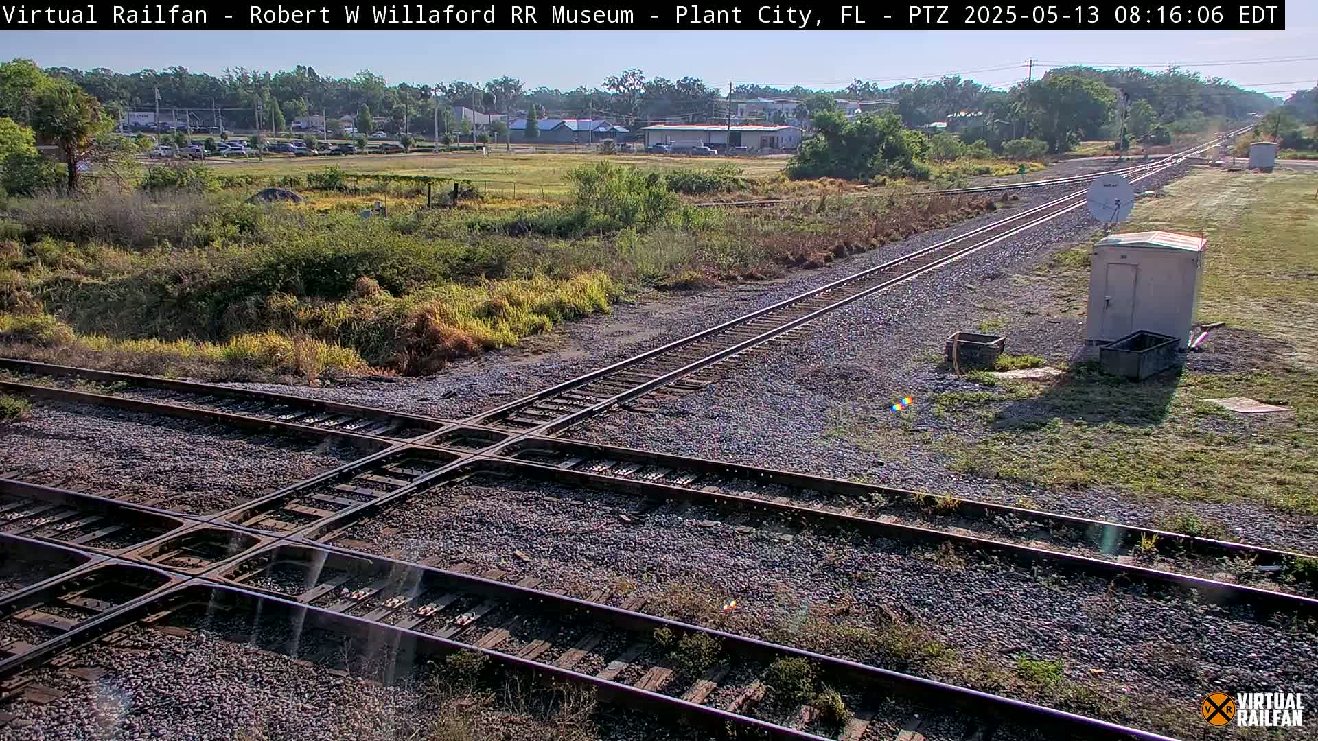 A sunny view of a railroad crossing with two sets of tracks intersecting, surrounded by scrubby vegetation and small utility structures.