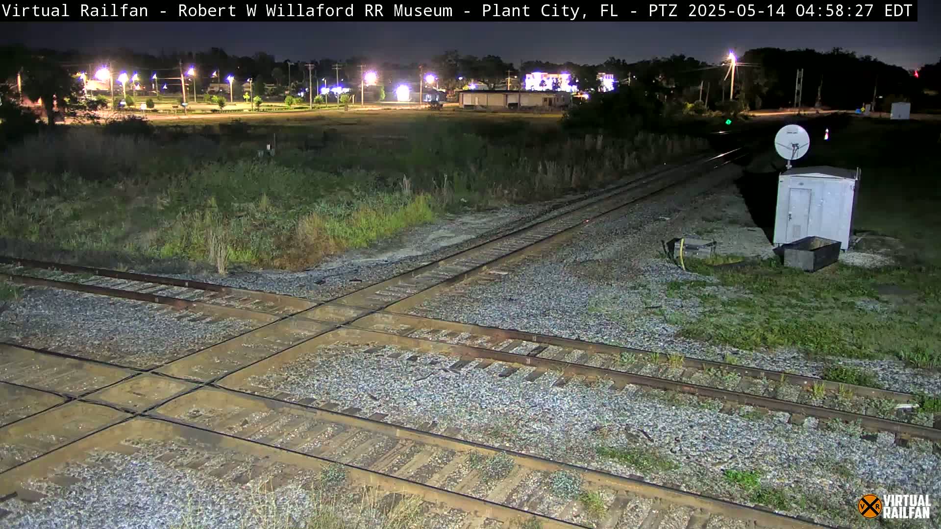 A nighttime view of a railroad crossing, with several tracks intersecting, surrounded by overgrown vegetation and a small building, under dark, clear skies.