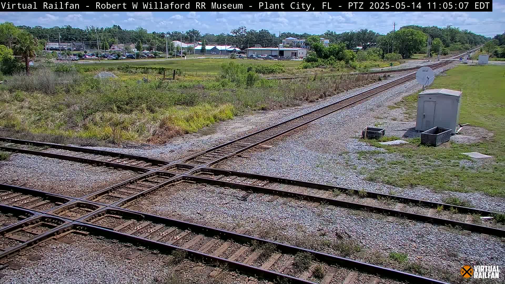 A railroad crossing with two sets of tracks is shown on a sunny day, with buildings and vegetation visible in the background.