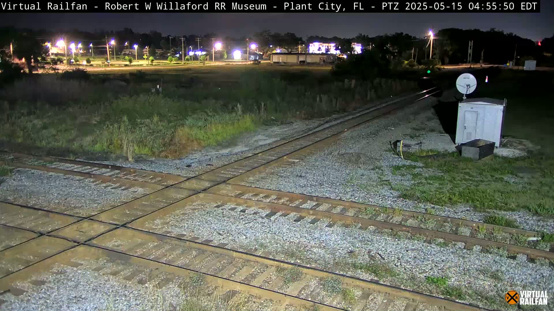 A nighttime view of a railroad crossing with a small building and satellite dish nearby, under possibly rainy conditions.