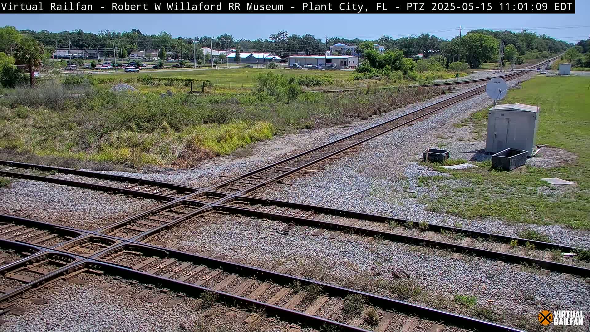 A sunny outdoor scene shows a railroad crossing with two sets of tracks intersecting, surrounded by sparse vegetation and a small building.