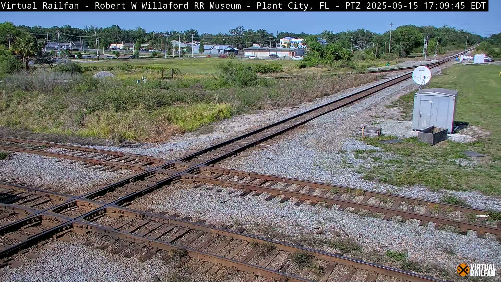 A sunny outdoor scene shows a railroad crossing with two sets of tracks intersecting, near a small building and satellite dish, with sparse vegetation and distant buildings visible in the background.