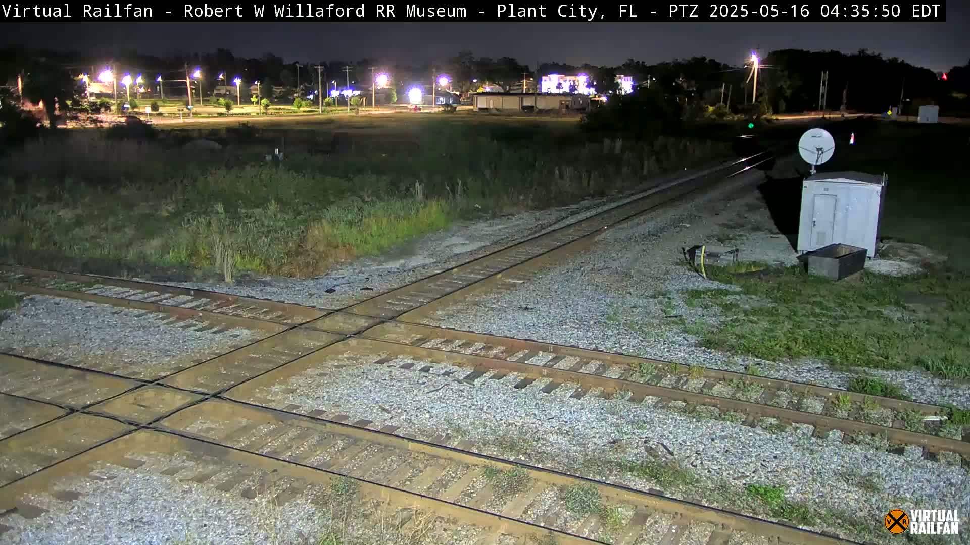A nighttime view of a railroad crossing with intersecting tracks, surrounded by vegetation and a small building, under dark conditions.
