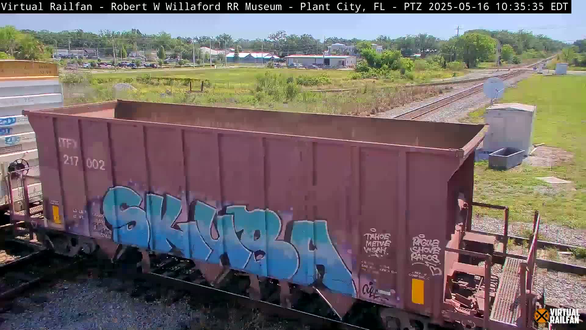 A rusty, graffiti-covered train car sits on railroad tracks on a sunny day, with other cars and distant buildings visible in the background.