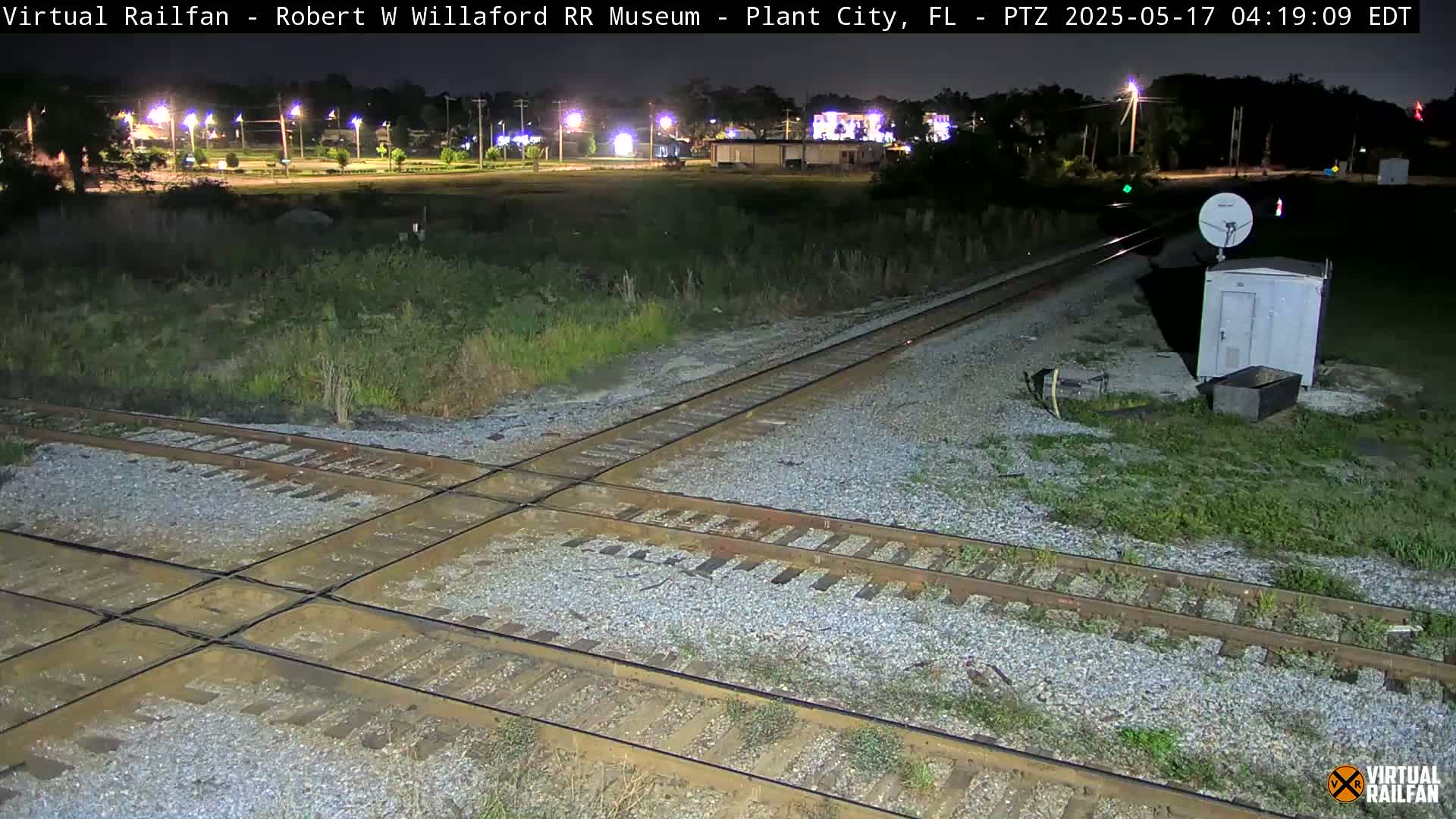 A nighttime view of a railroad crossing with a small building and satellite dish nearby, under dark skies.