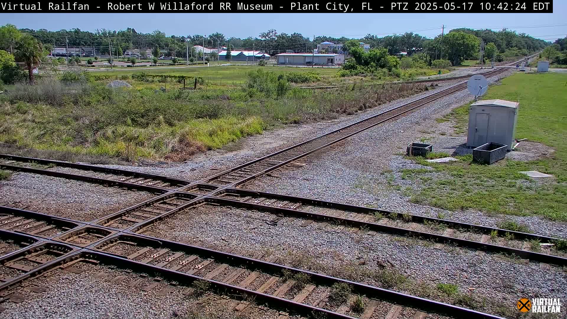Railroad tracks intersect at a crossing, under a sunny sky, with vegetation and a small building visible in the background.