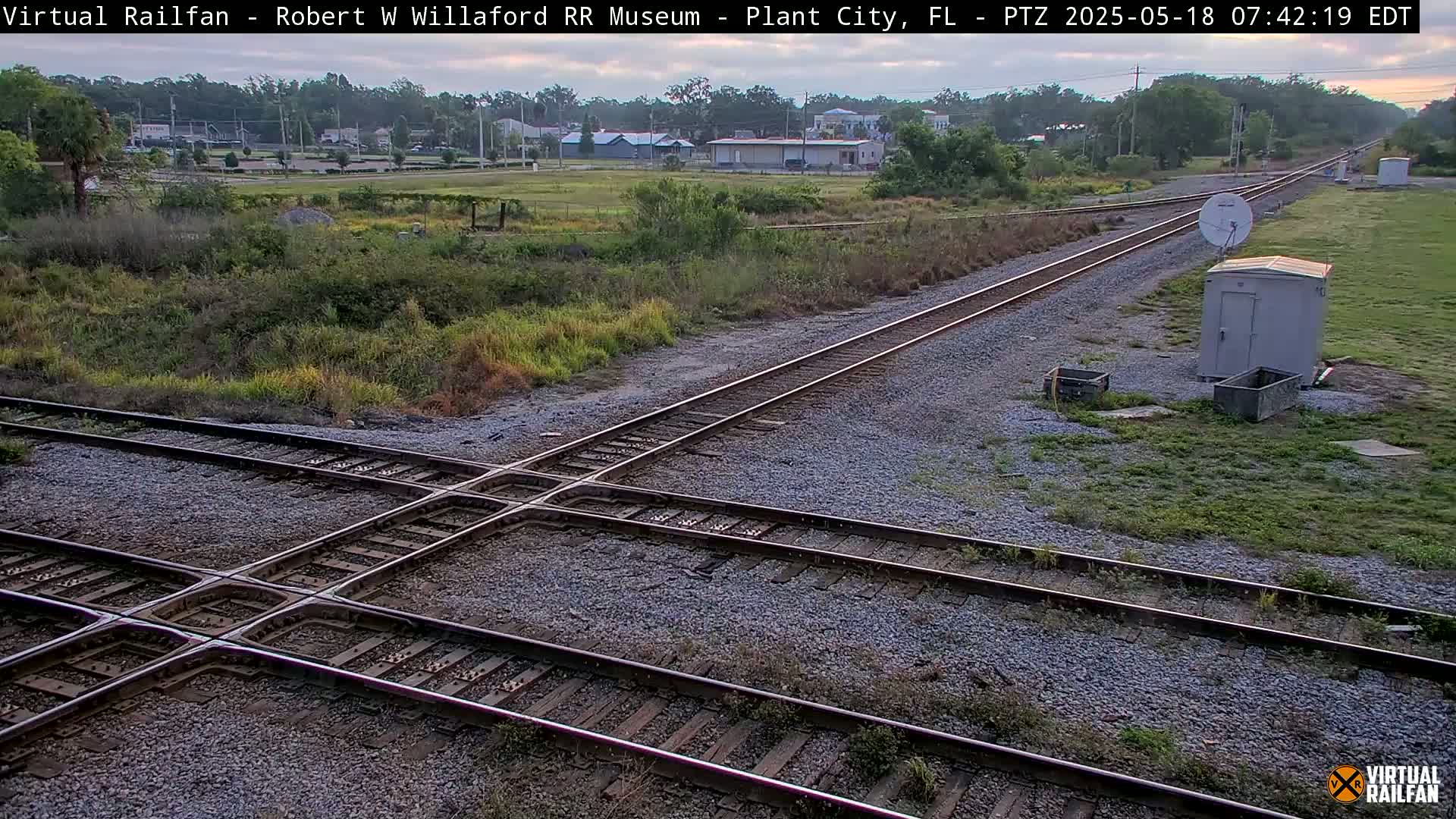 A railroad crossing with two sets of tracks intersects at a slight angle, under a partly cloudy sky, in a grassy area near some buildings.