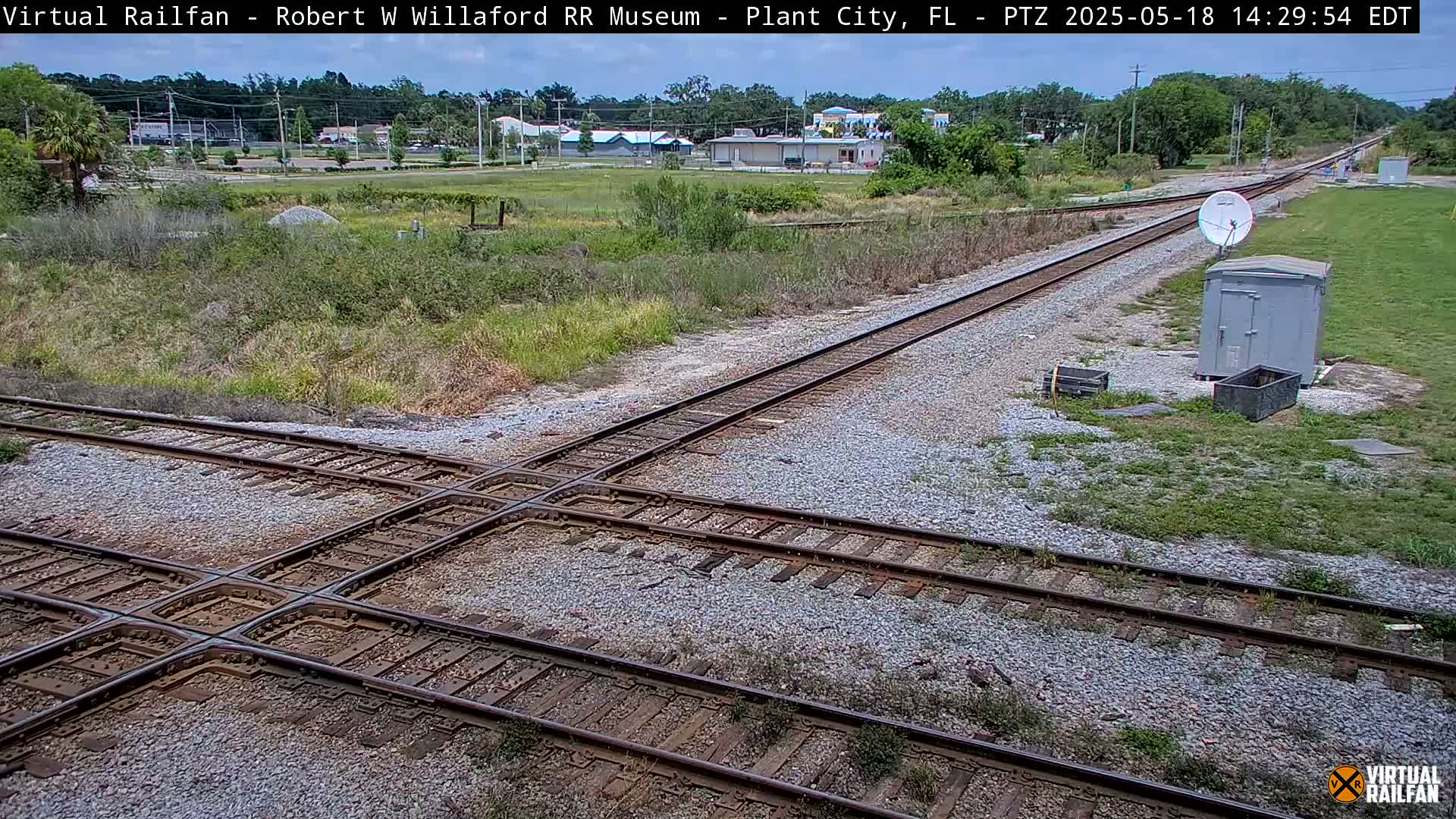 A railway crossing with two sets of tracks intersecting, situated in a grassy area near buildings under a partly sunny sky.