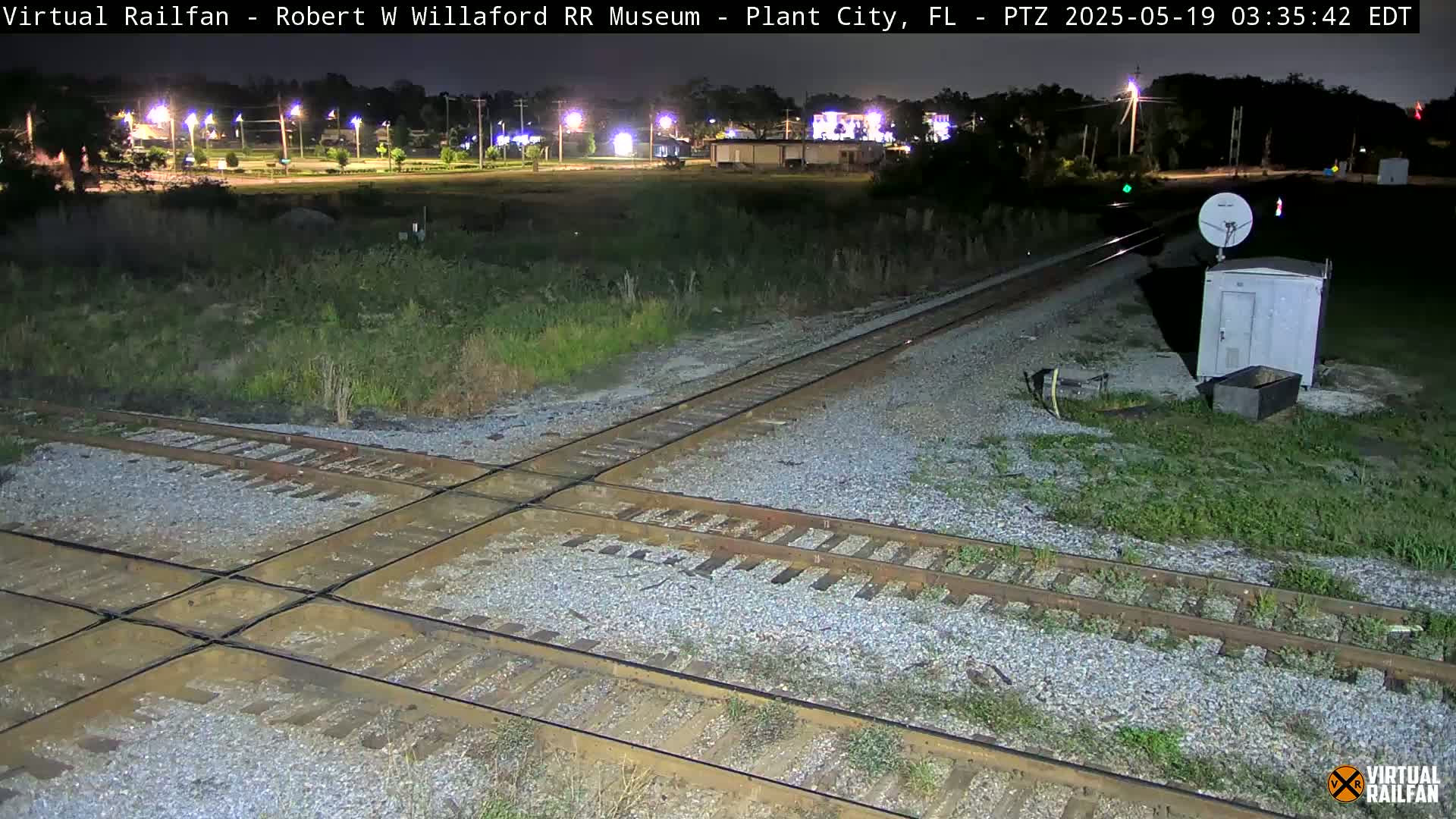 A nighttime view of a railroad crossing with a small building and satellite dish nearby.
