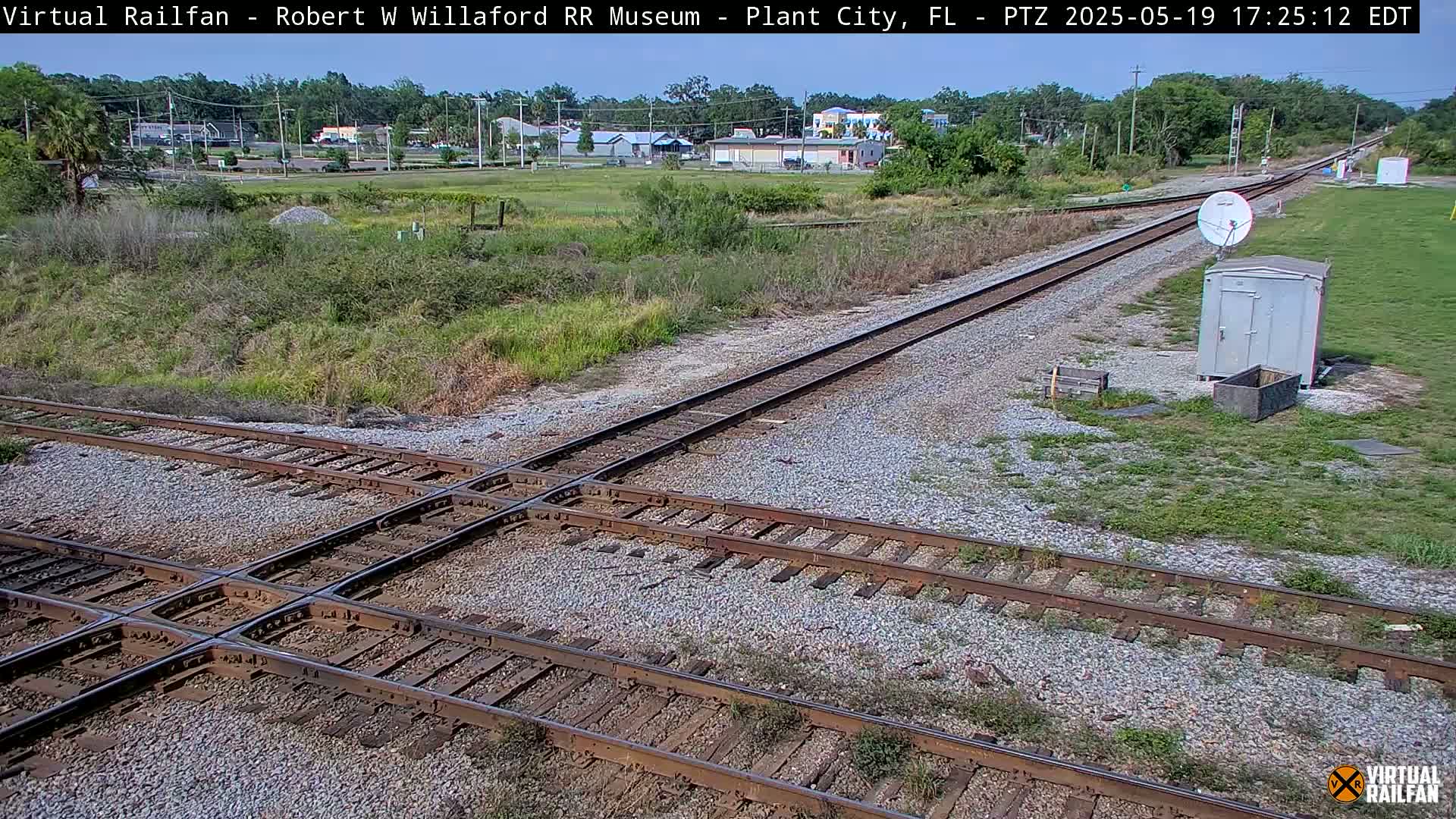 A high-angle view shows a railroad crossing under a partly sunny sky, with some sparse vegetation and a small building in the distance.