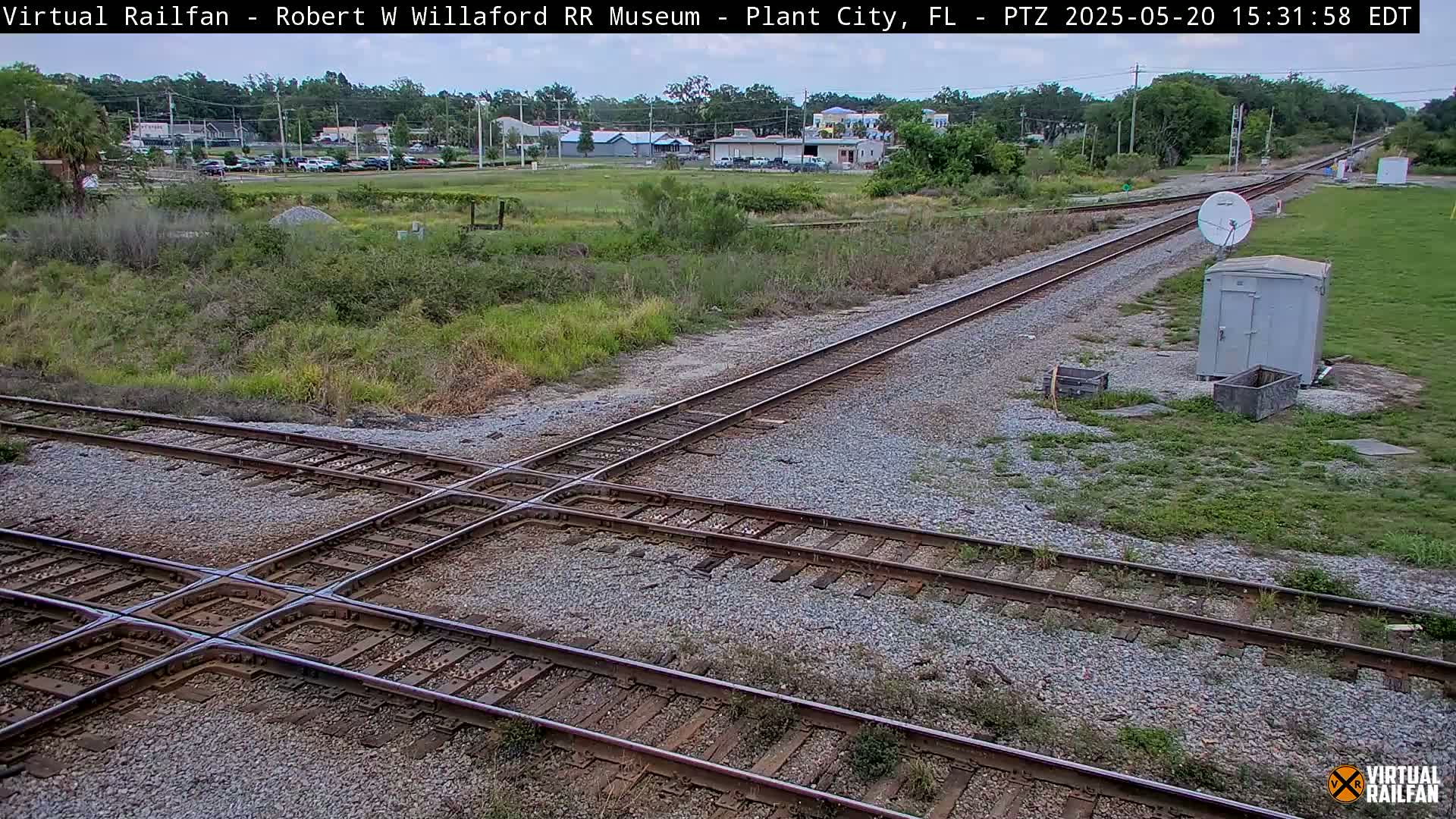 A train crossing intersects two railway lines, amidst overgrown vegetation and a small building, under a partly cloudy sky.