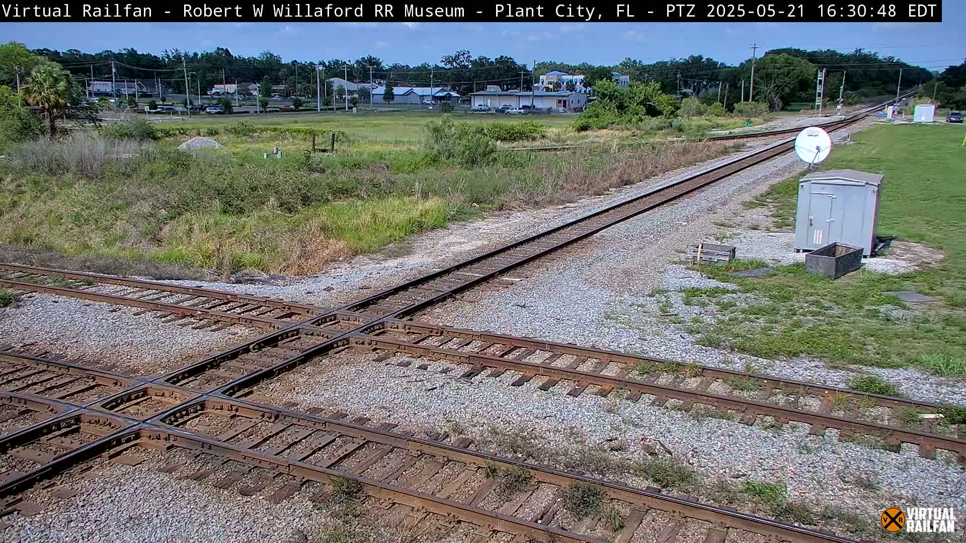 A high-angle view of a railroad crossing on a sunny day, showing two sets of tracks intersecting, with sparse vegetation and a small building visible in the distance.