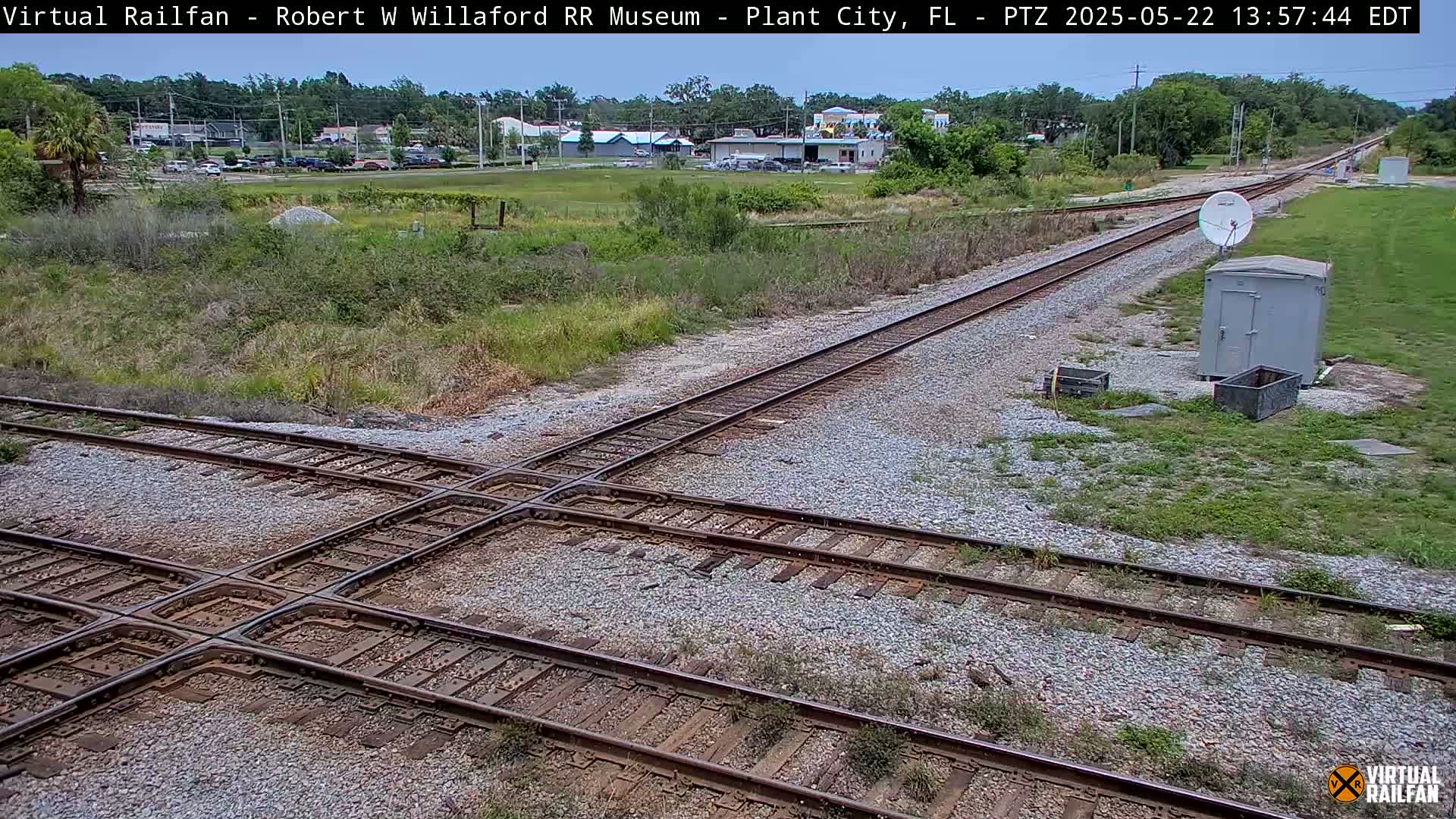 A railroad crossing with two sets of tracks intersecting, surrounded by scrubby vegetation under a partly sunny sky.