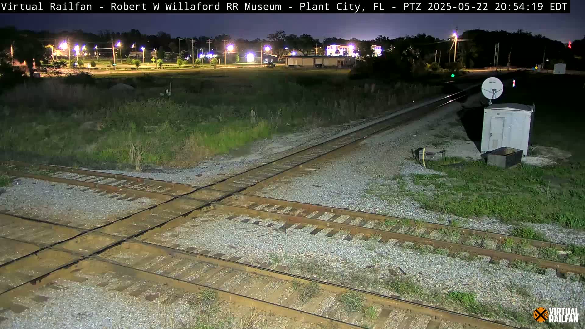 A nighttime view of a railroad crossing with two sets of tracks intersecting, a small utility building nearby, and a sparsely vegetated area surrounding it.