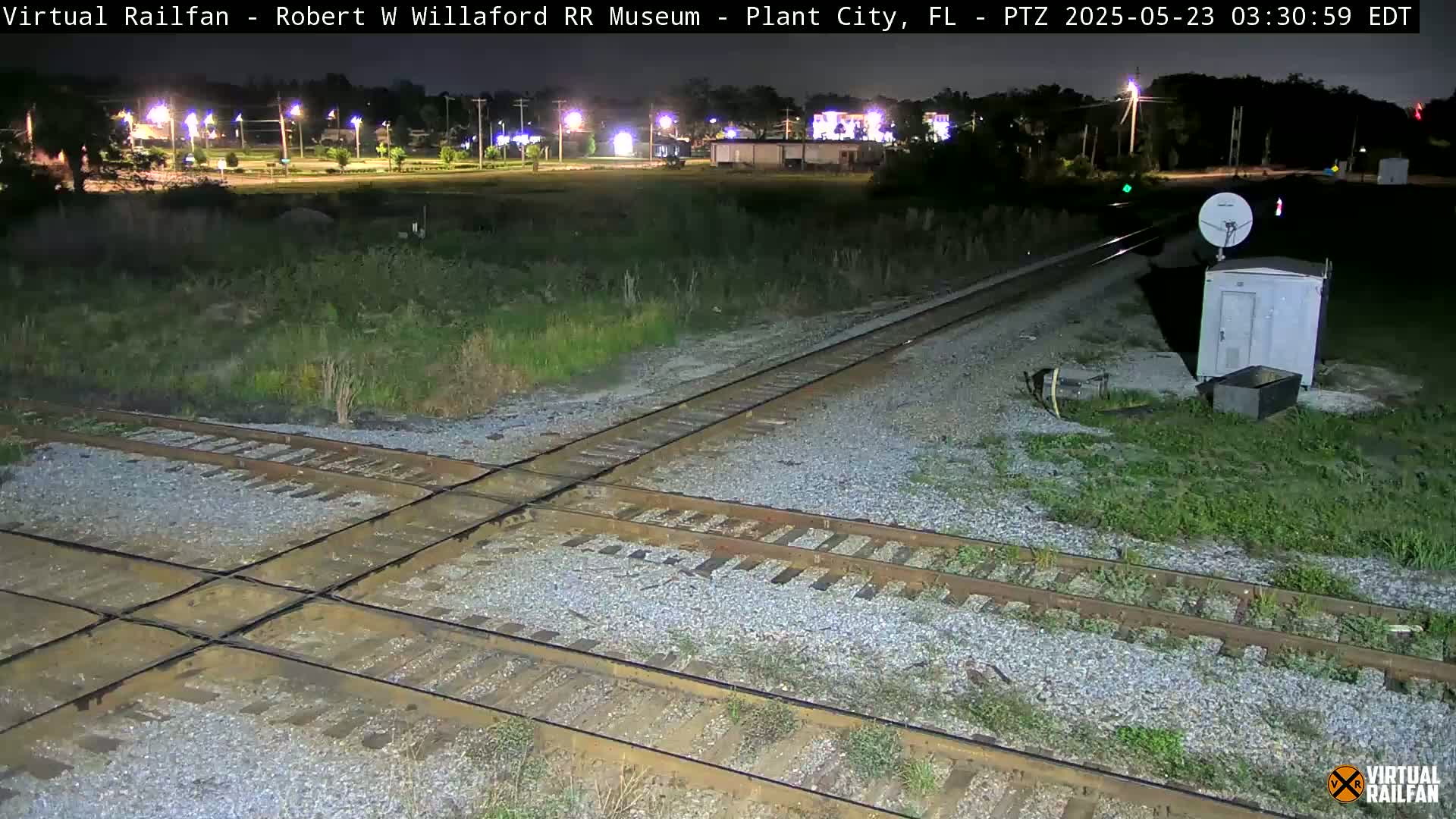 A nighttime view of a railroad crossing with a small building and satellite dish nearby, under clear skies.