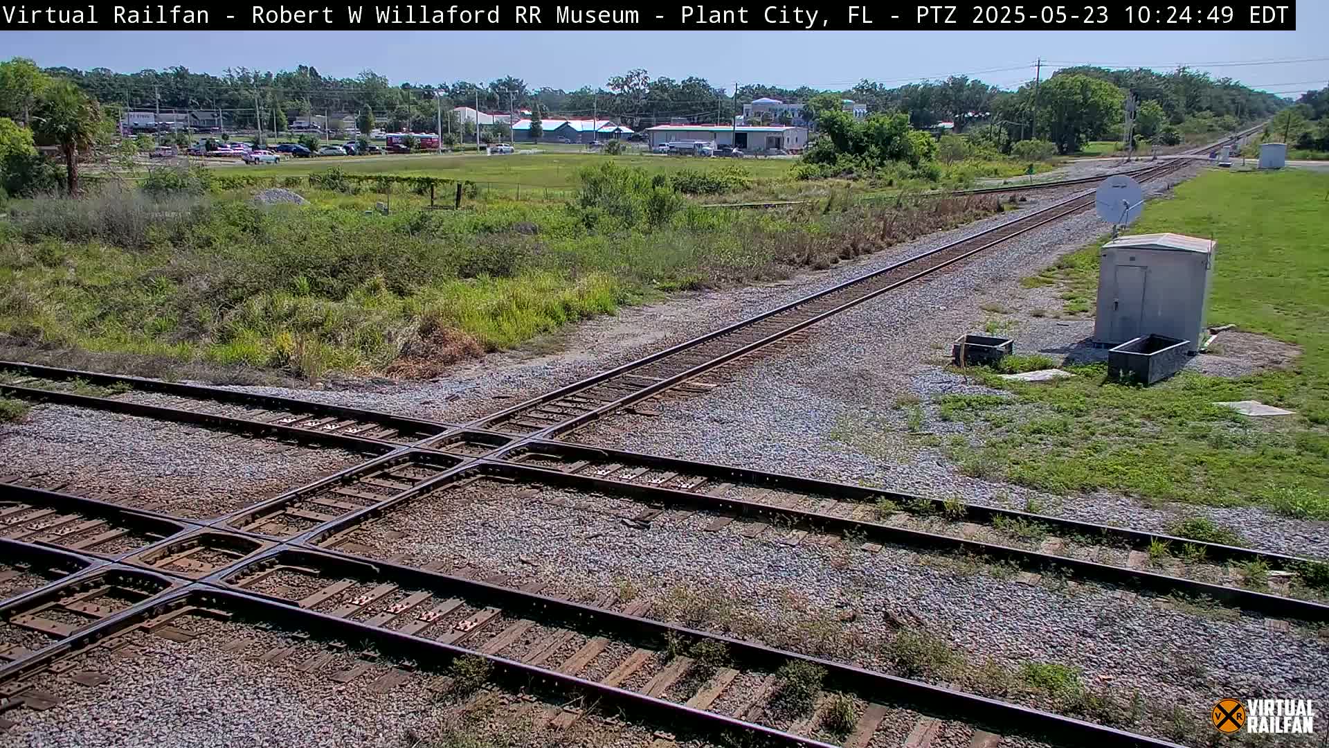 A sunny outdoor scene shows a railroad crossing with several tracks intersecting, surrounded by grassy areas and some small buildings in the distance.