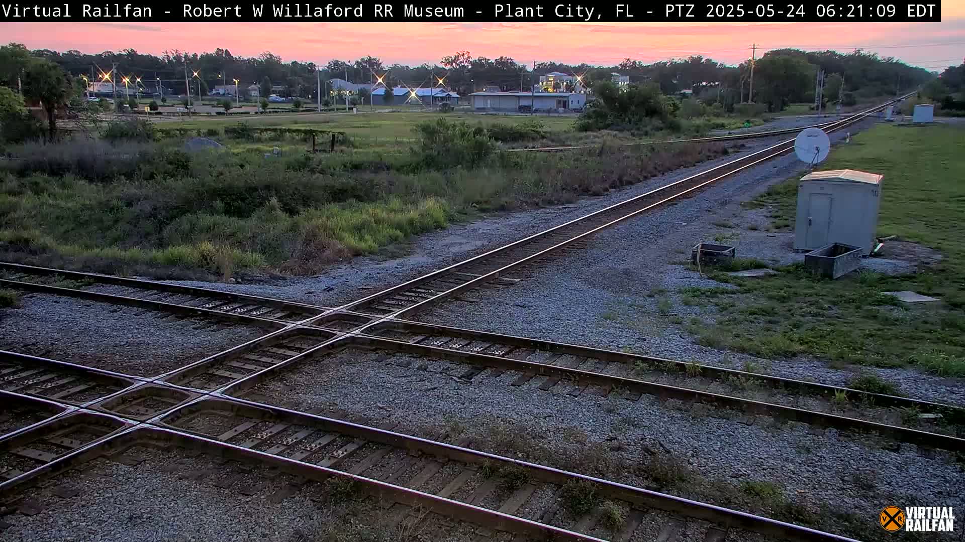 A railroad crossing at dawn, with train tracks intersecting against a backdrop of low vegetation and distant buildings.
