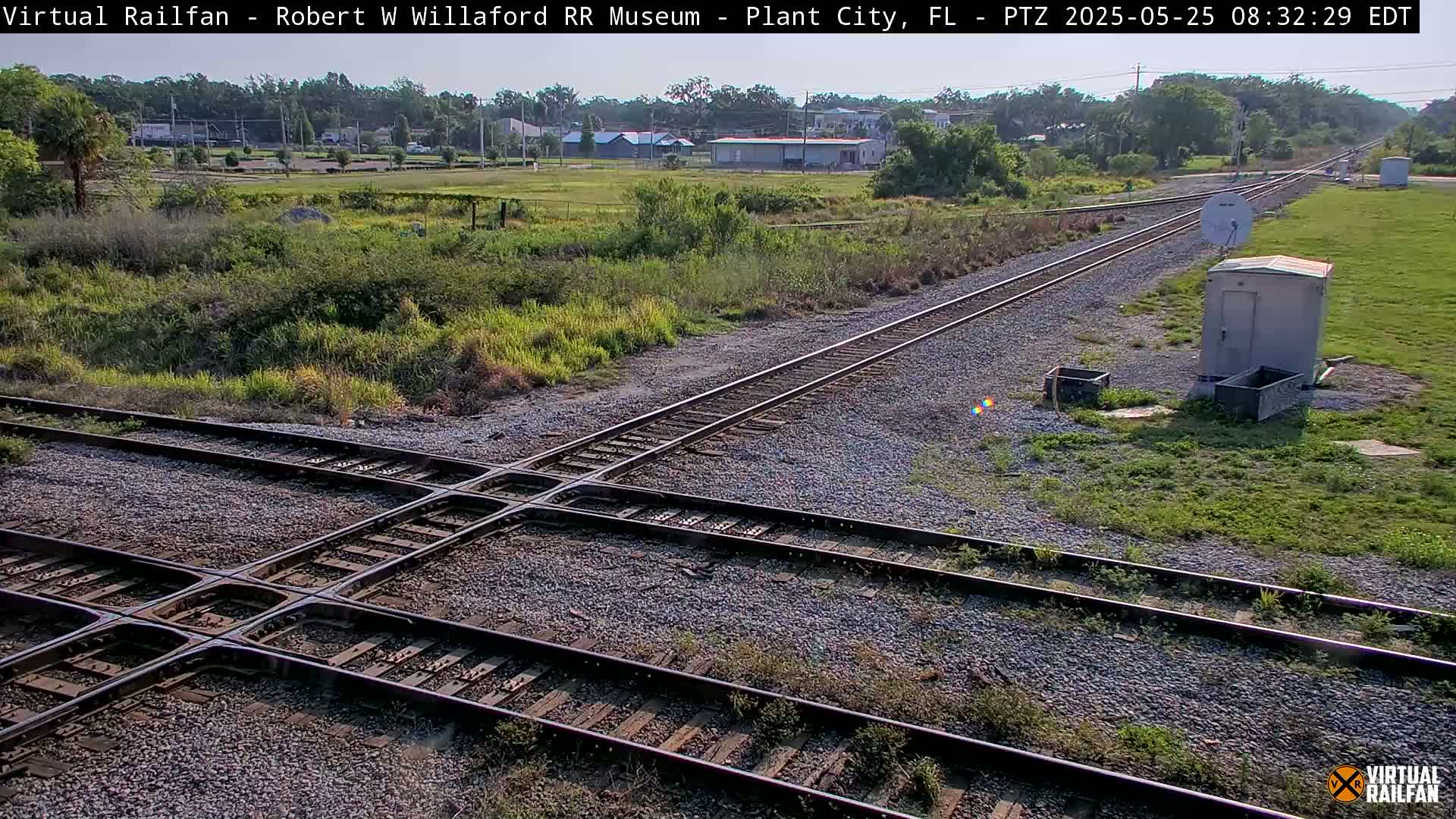 A high-angle, long shot of a railroad crossing on a sunny day, with sparse vegetation surrounding the tracks and a small building nearby.