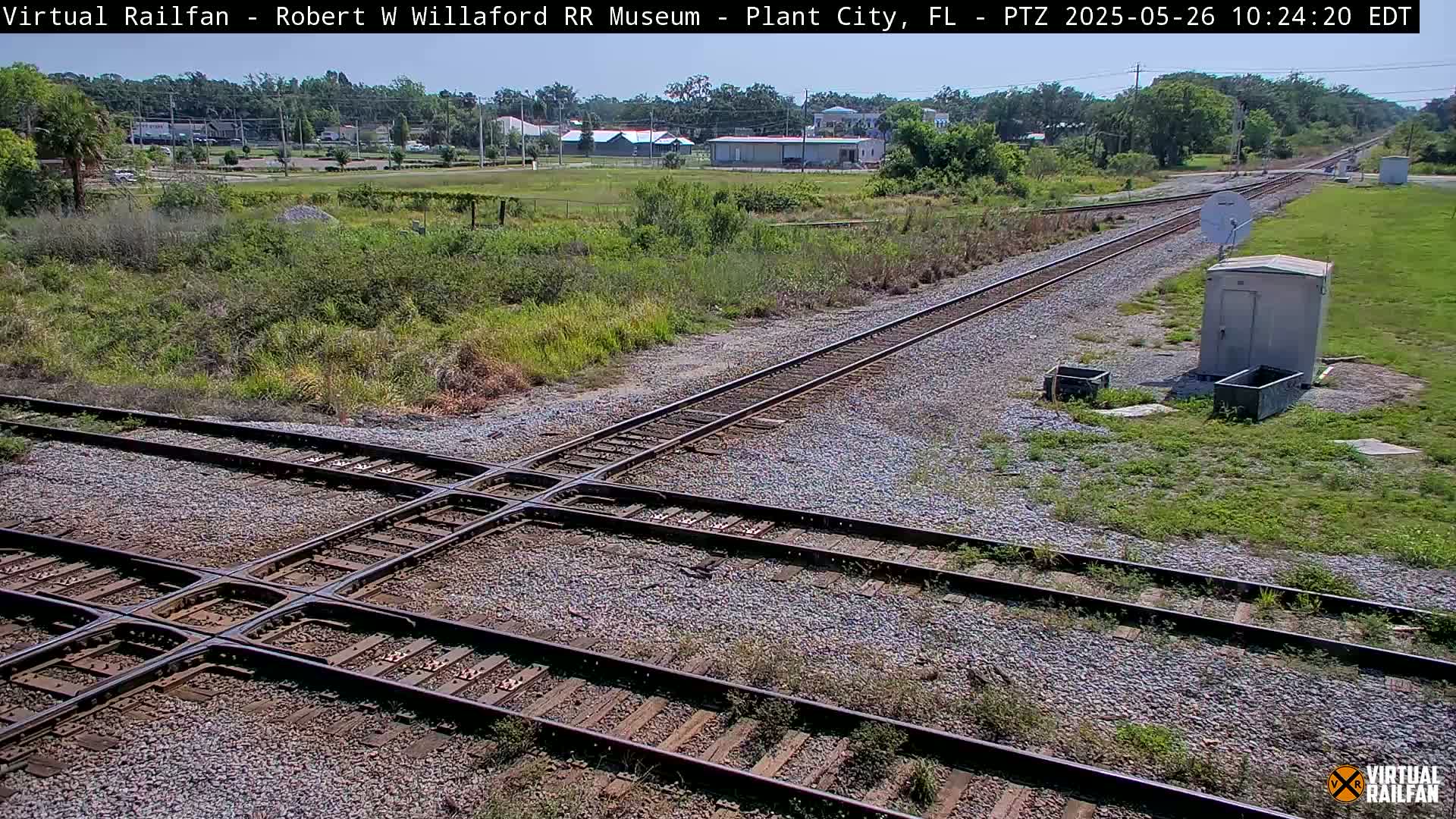 A sunny outdoor view shows a railroad crossing with two sets of tracks intersecting, surrounded by patchy grass and a small utility building.