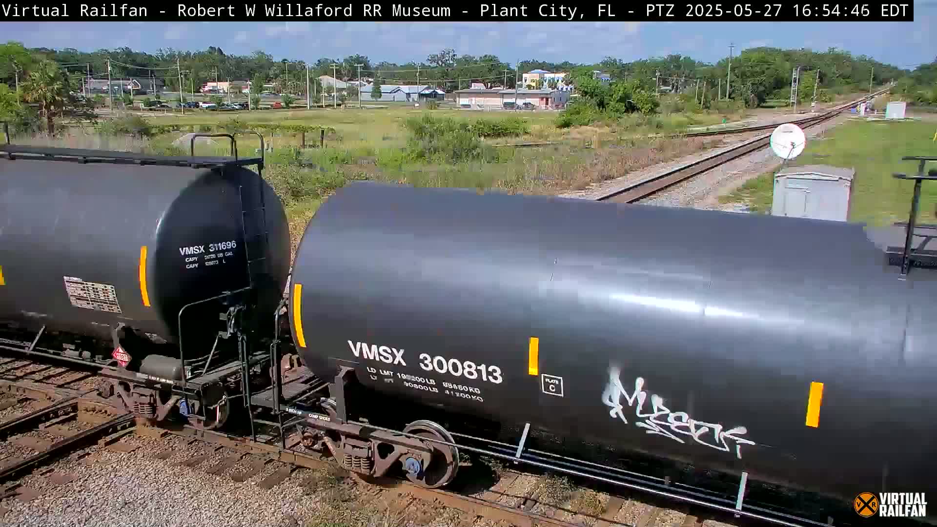 Two dark-gray tanker cars are on a railway track, with a grassy area and buildings in the background on a sunny day.