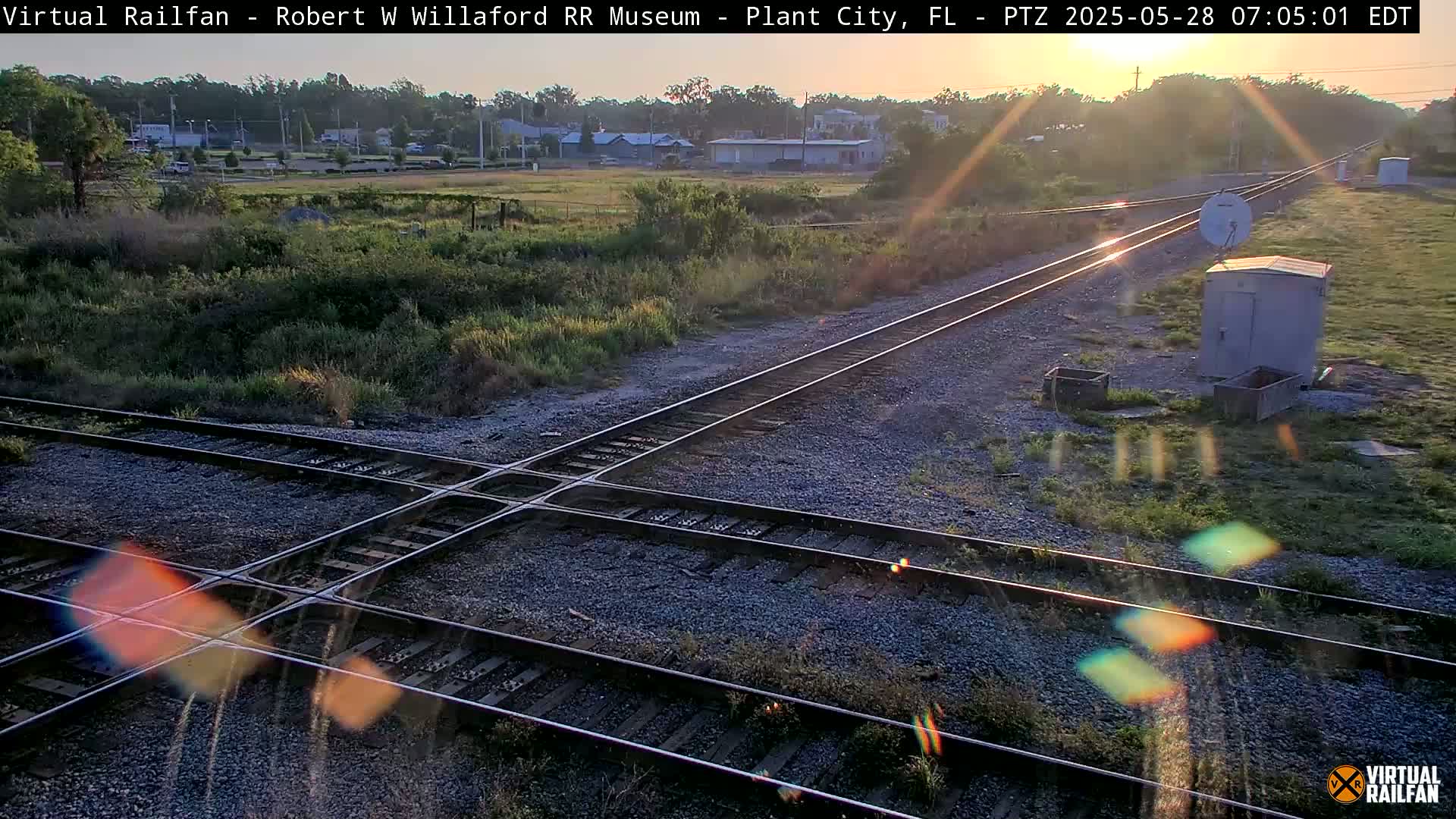 A high-angle, long shot of a railroad crossing at sunrise, showing two sets of train tracks intersecting, surrounded by scrub vegetation and a small building.