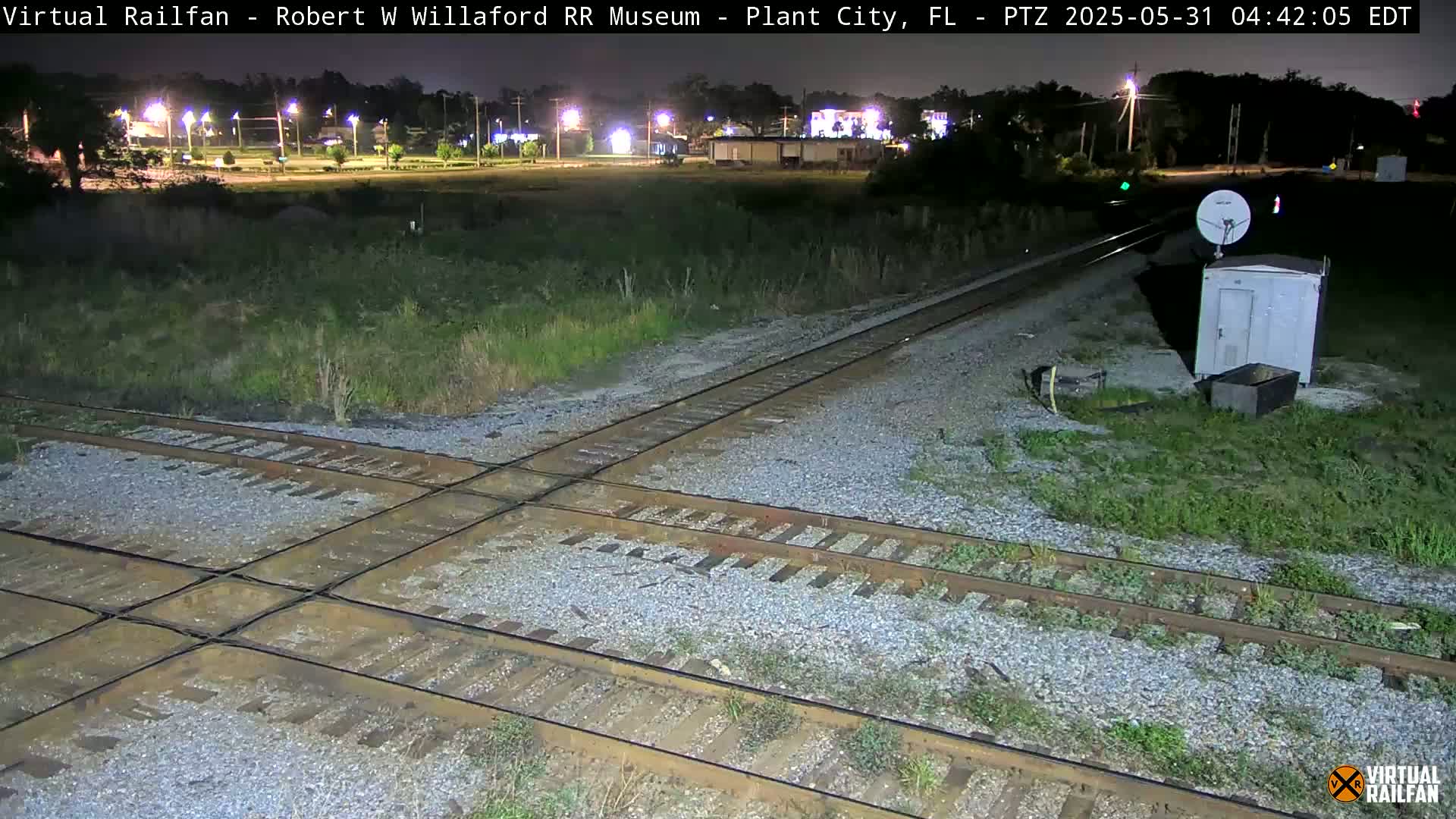 A nighttime view of a railroad crossing with a small building and satellite dish nearby.