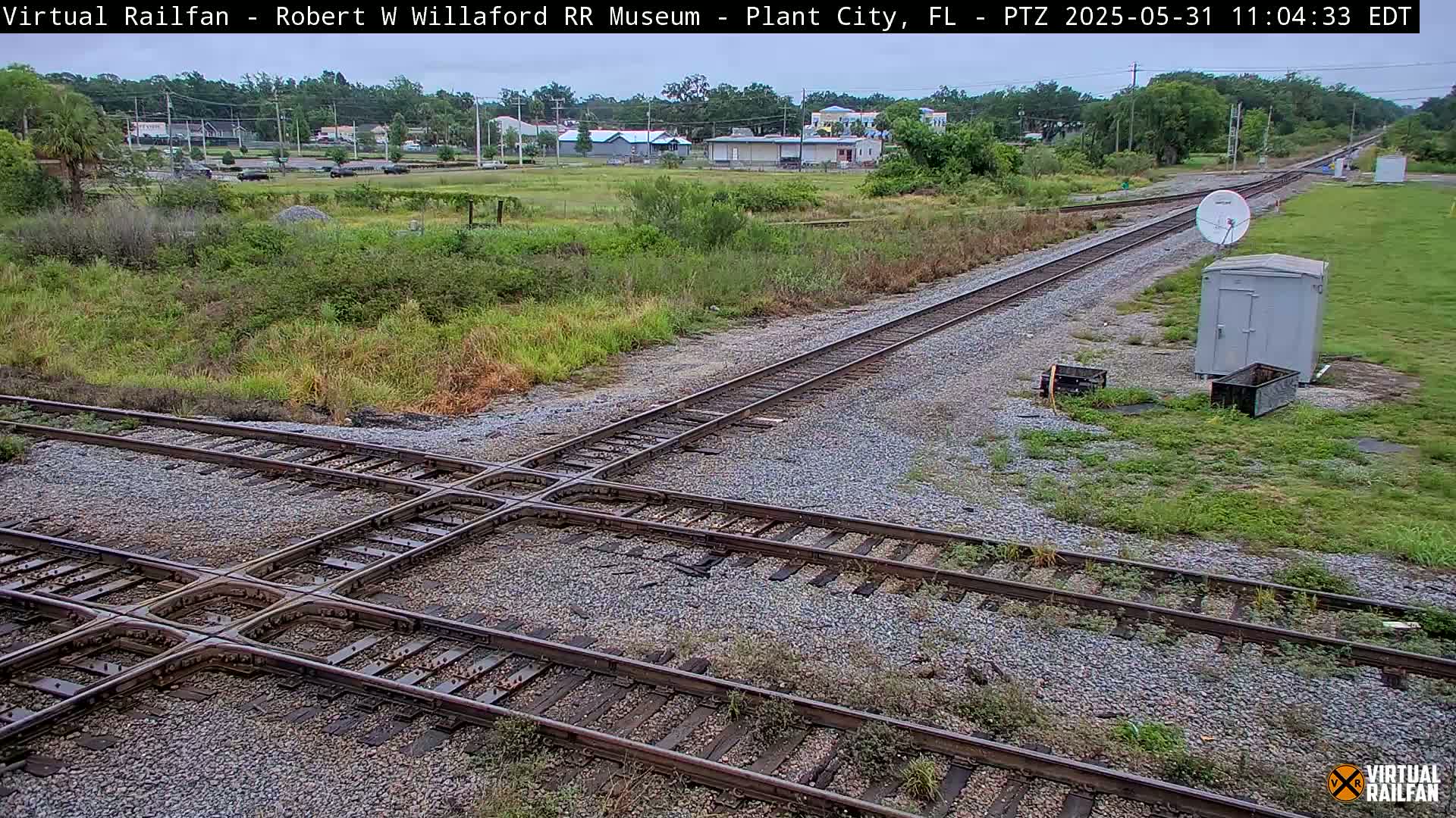 A railroad crossing with intersecting tracks is shown on a partly cloudy day, surrounded by overgrown vegetation and a small building.