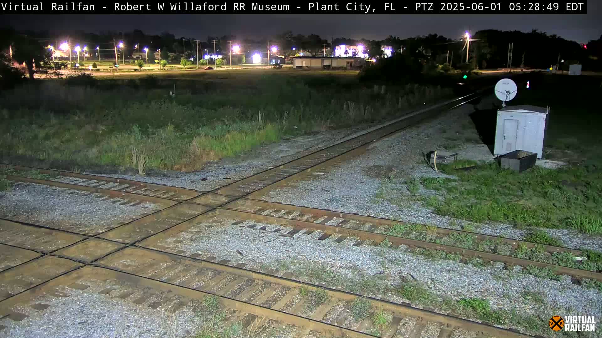 A nighttime view of a railroad crossing with intersecting tracks, surrounded by overgrown vegetation and a small building with a satellite dish nearby.