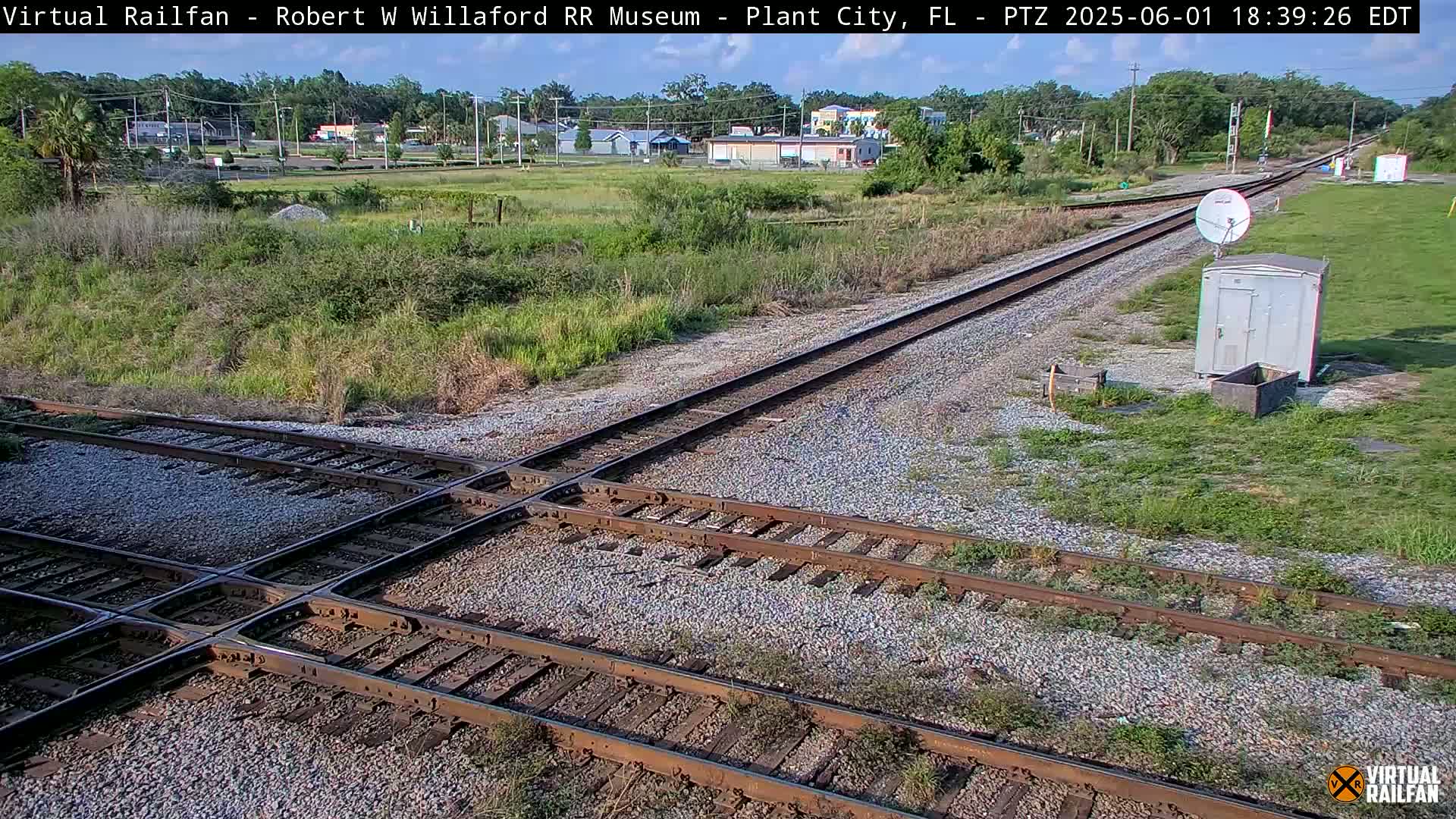 A railroad crossing with two sets of tracks intersecting, surrounded by scrub vegetation and a small building, under a sunny sky.