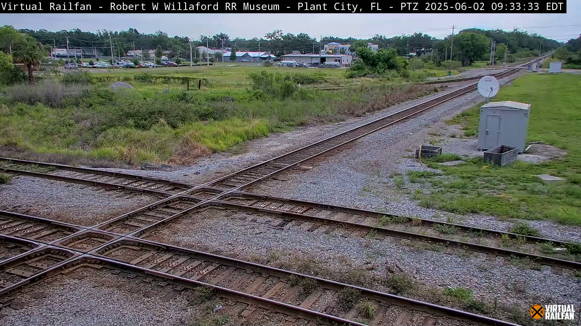 A railroad crossing with several tracks intersecting, under partly cloudy skies, surrounded by overgrown vegetation and a small building.