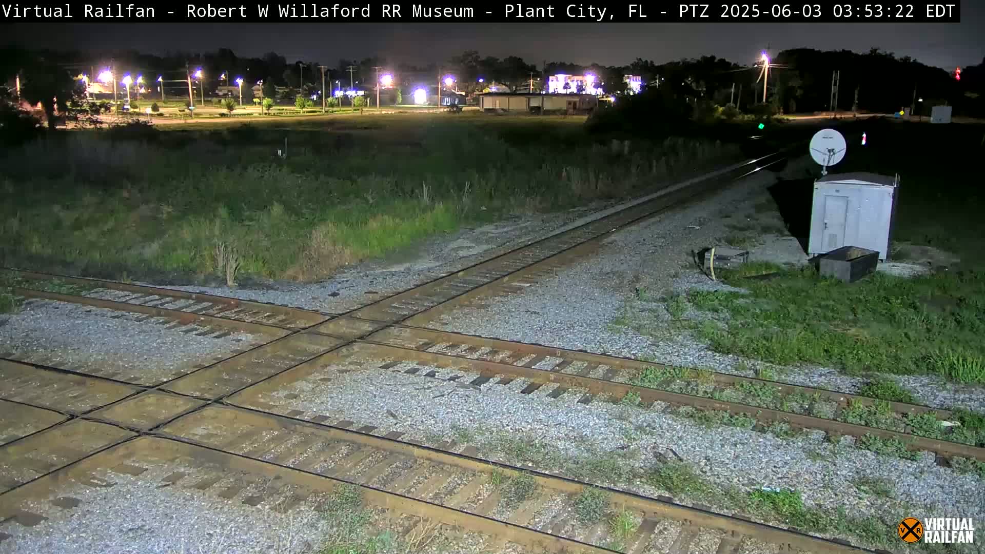 A nighttime view of a railroad crossing with a small building and satellite dish, under a clear sky.