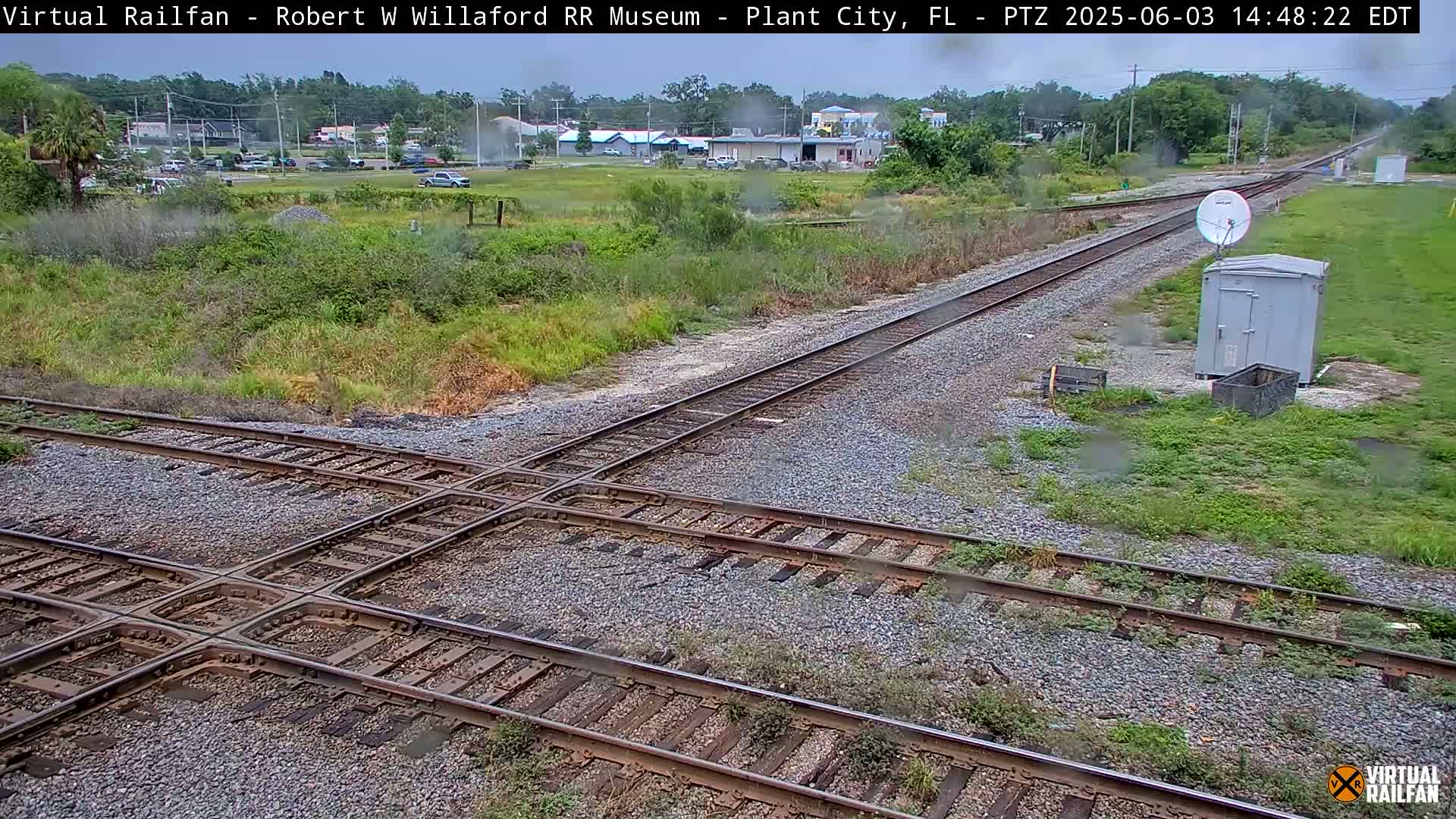 A railroad crossing with two sets of tracks intersecting, situated in a grassy area with some light rain, and a small building and satellite dish nearby.