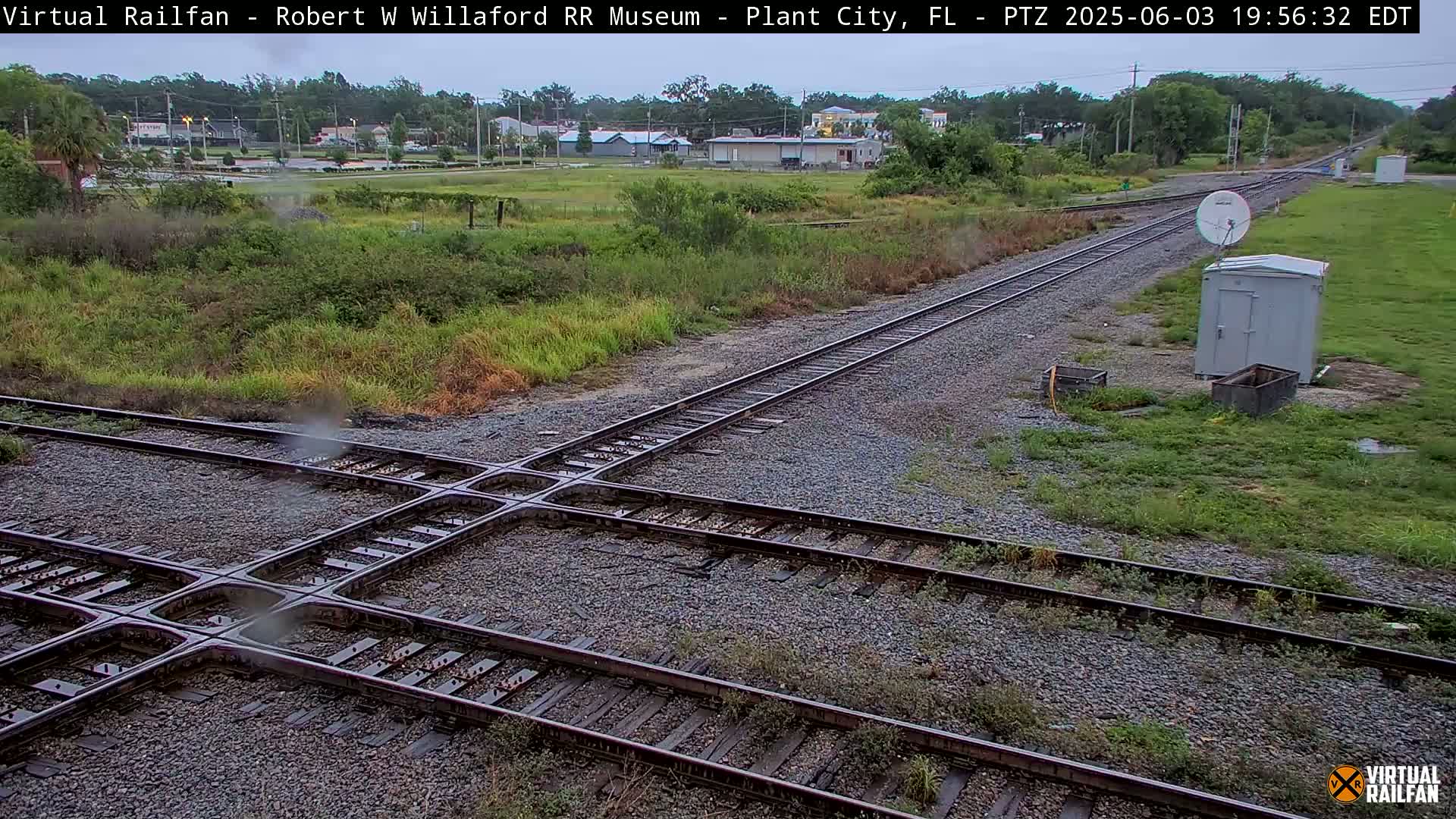 A railroad crossing with two sets of tracks intersecting, surrounded by overgrown vegetation and a small building with a satellite dish nearby, under overcast skies.