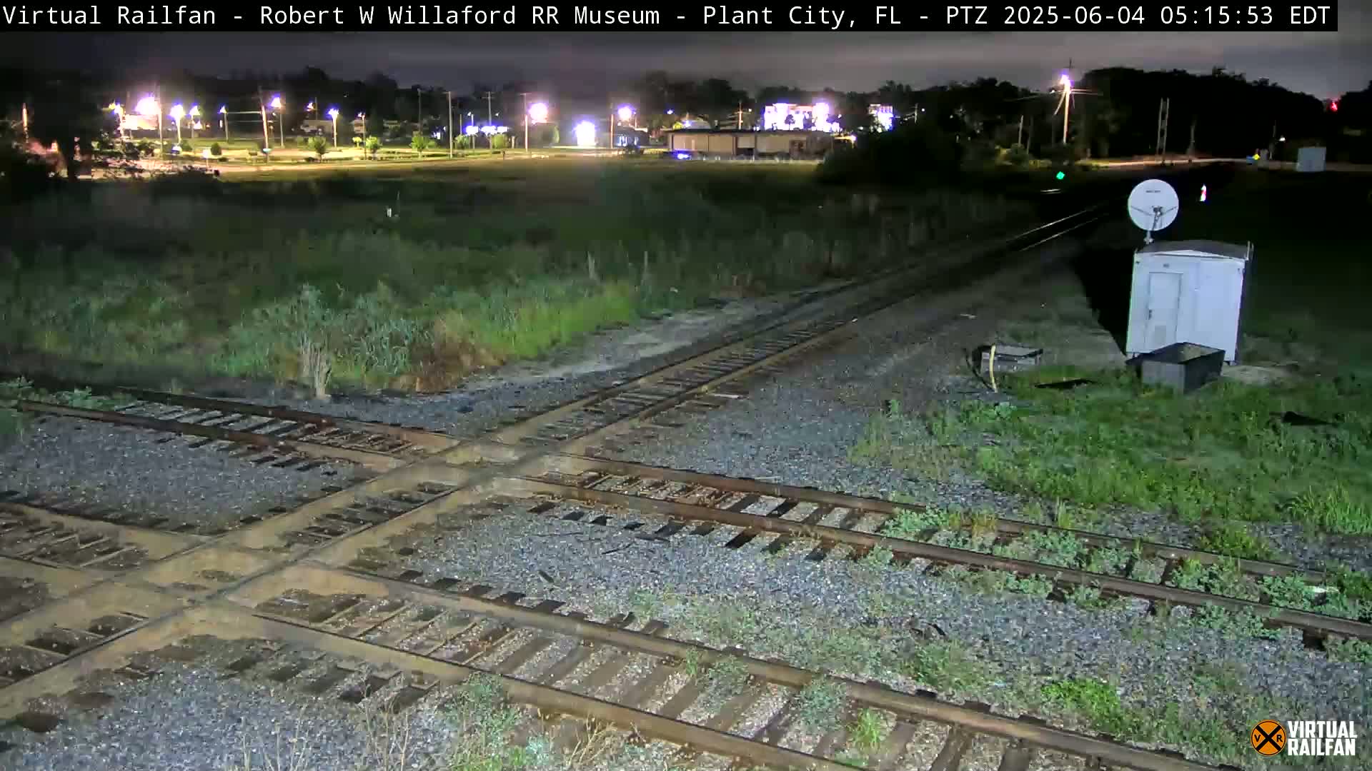 A nighttime view of a railroad crossing with intersecting tracks, surrounded by overgrown vegetation, under a dark sky.