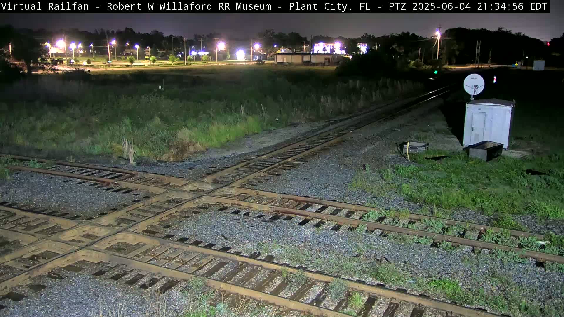 A nighttime view of a railroad crossing with a small building and satellite dish near the tracks, under partly cloudy skies.