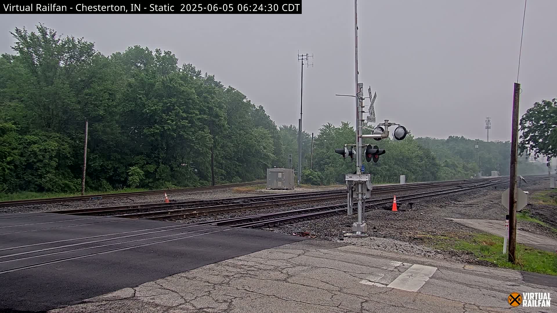 A railroad crossing with a signal and multiple tracks is seen on an overcast day, surrounded by lush green trees.