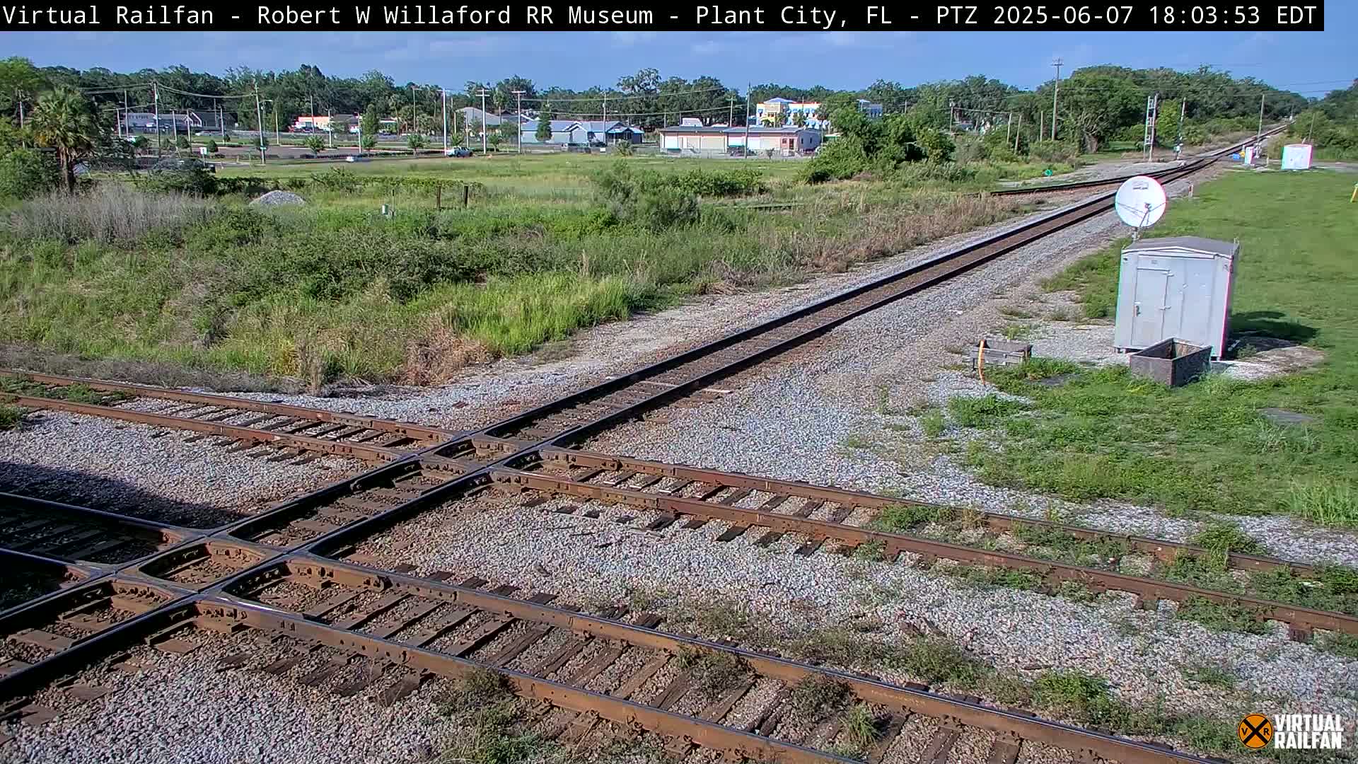 A high-angle, sunny view of a railroad crossing with intersecting tracks, surrounded by grassy areas and a small building.