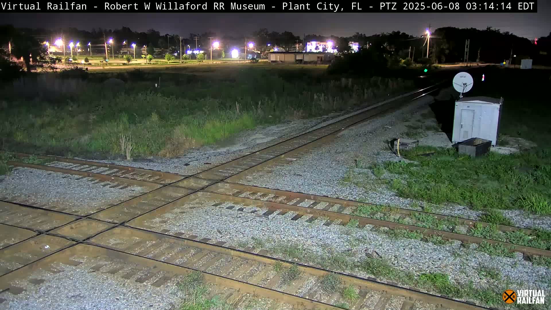 A nighttime view of a railroad crossing with a small building and satellite dish nearby, under clear skies.