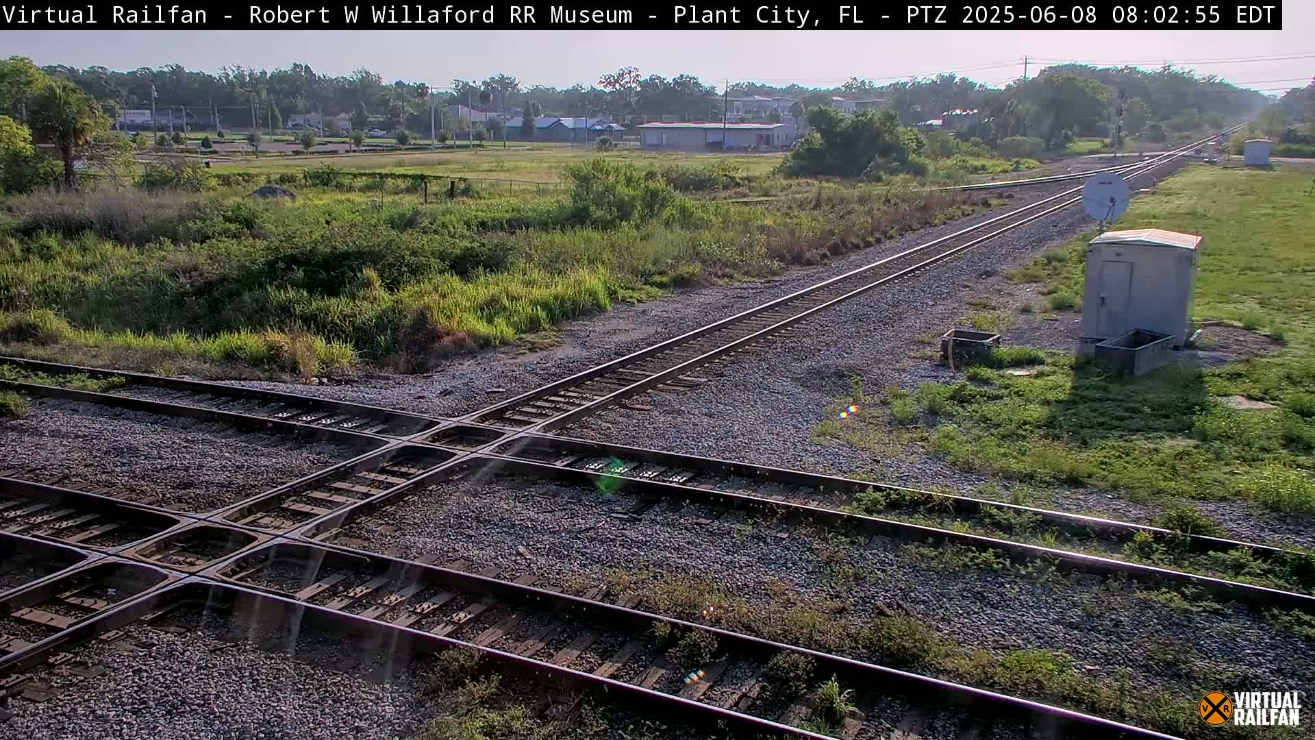 A sunny day shows a railroad crossing with two sets of tracks intersecting, surrounded by grassy fields and a small building.