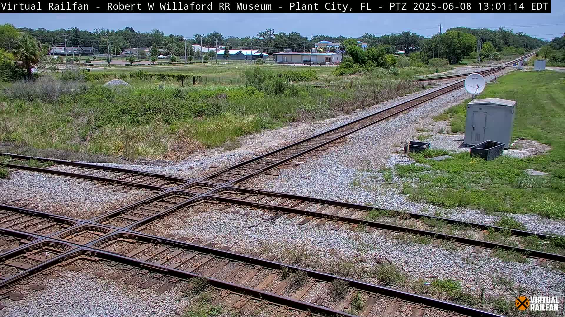 A railroad crossing with intersecting tracks sits amidst grassy fields on a sunny day, with buildings visible in the distance.