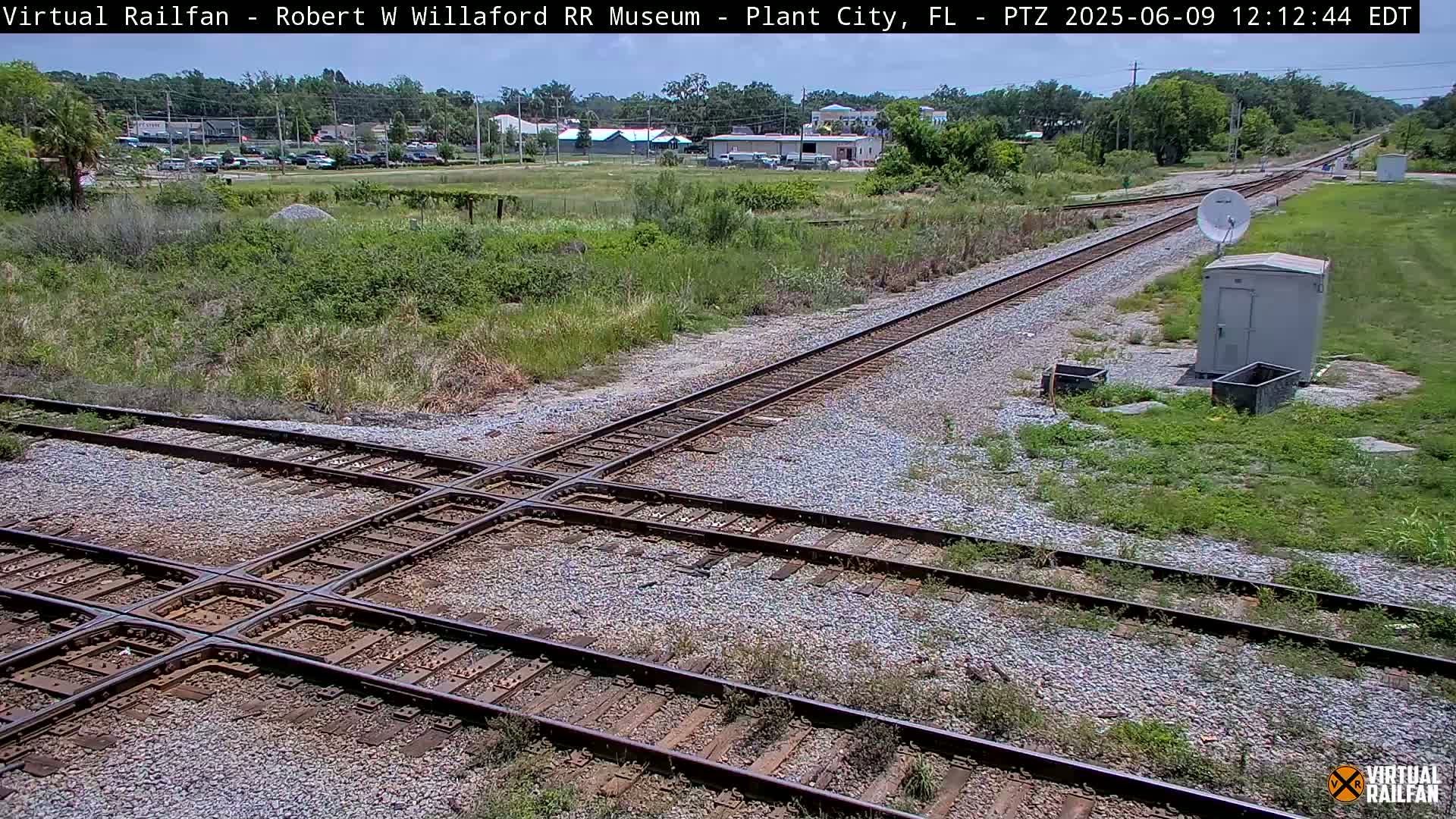 A sunny overhead view of a railroad crossing with two sets of tracks intersecting, surrounded by scrubby vegetation and a small building in the background.