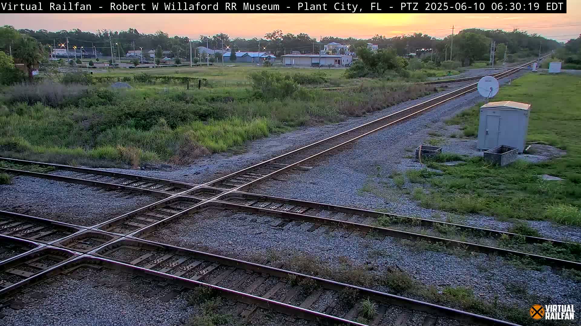 A high-angle, long shot shows a railroad crossing at sunset with a small building and satellite dish nearby.